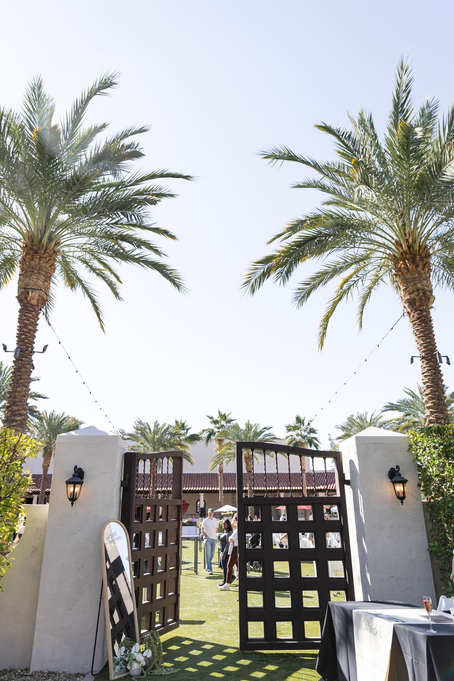 Open wooden gates flanked by tall palm trees lead to an outdoor wedding venue with string lights on a sunny day.