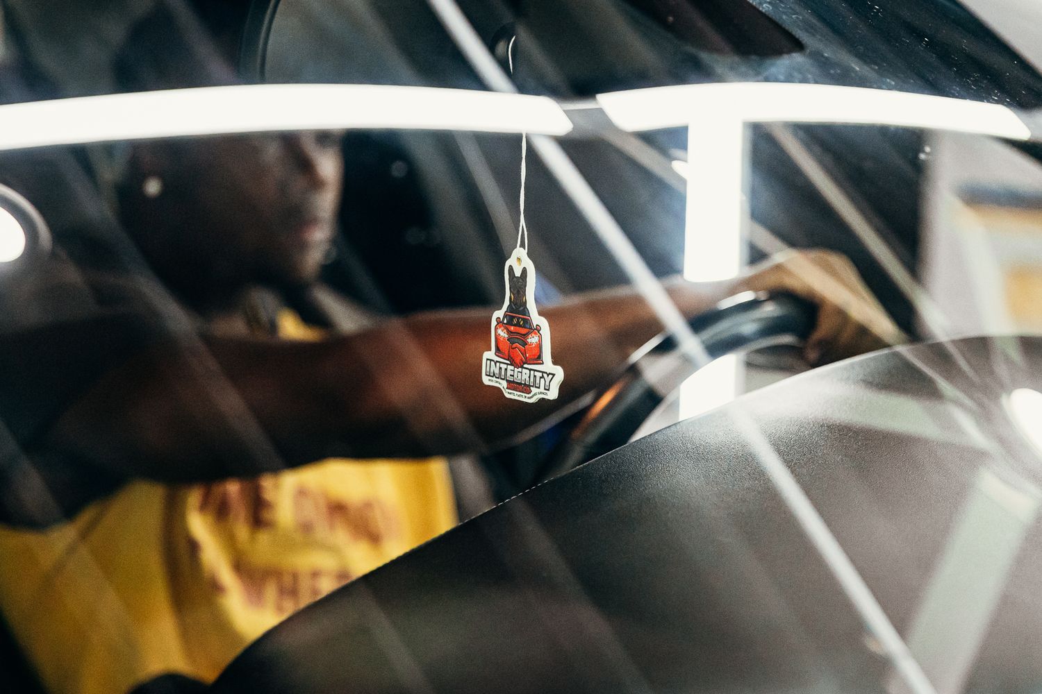 Person sitting in a car with a hanging air freshener ornament visible through the windshield.