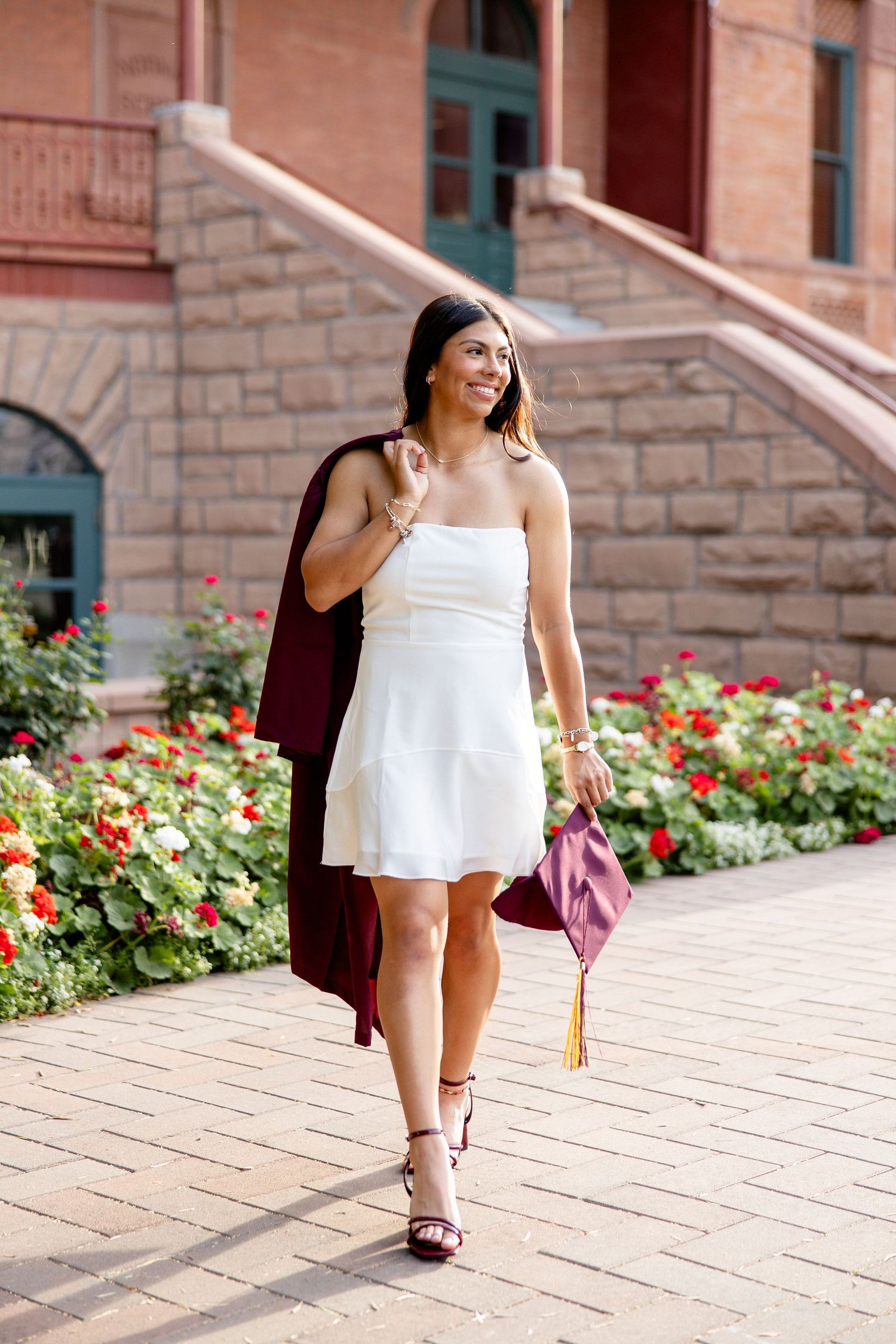 A woman in a white strapless dress walks past red flowers, carrying a maroon graduation cap and gown.