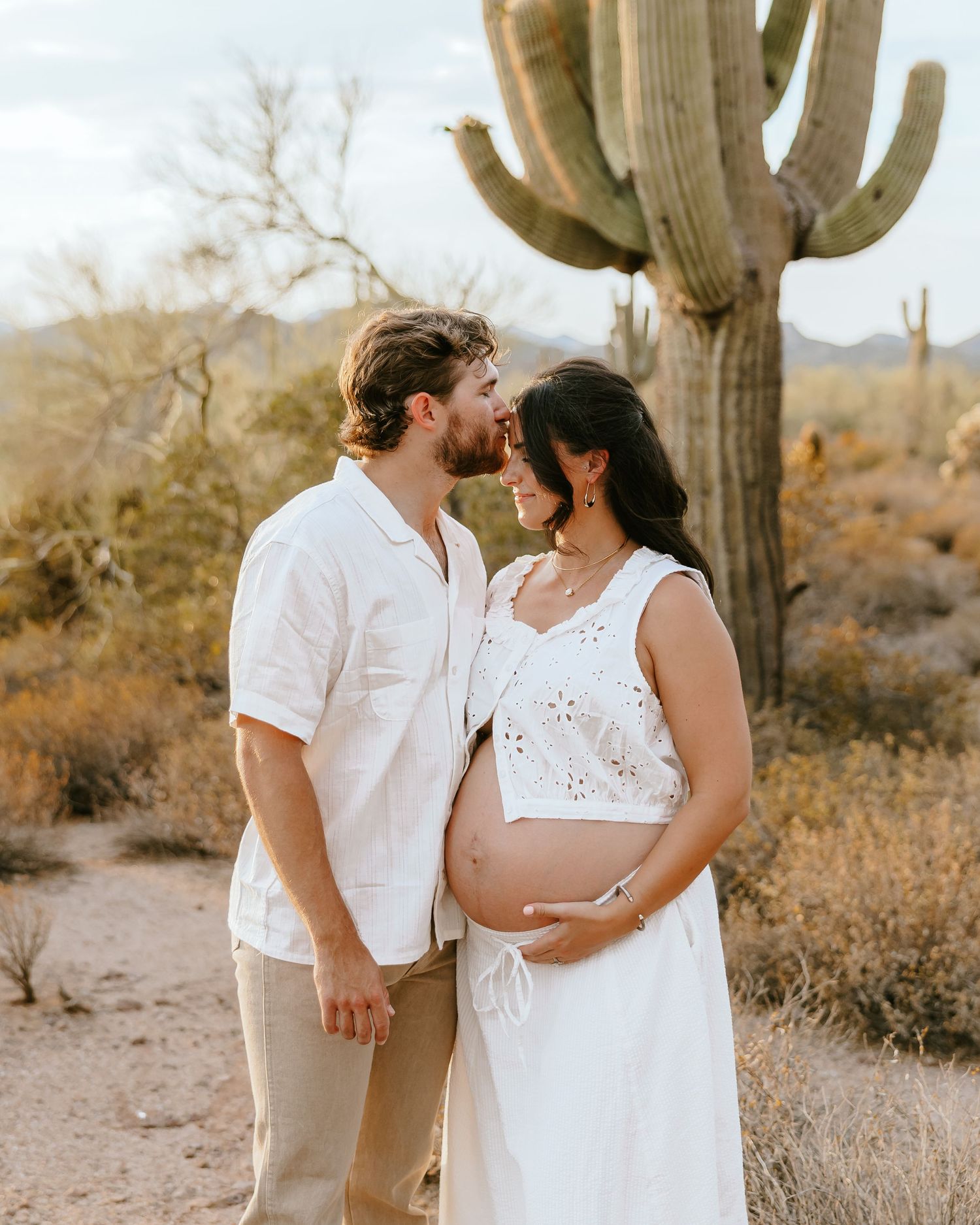 Couple sharing a tender moment in desert with saguaro cactus, pregnant woman in white dress cradling baby bump.