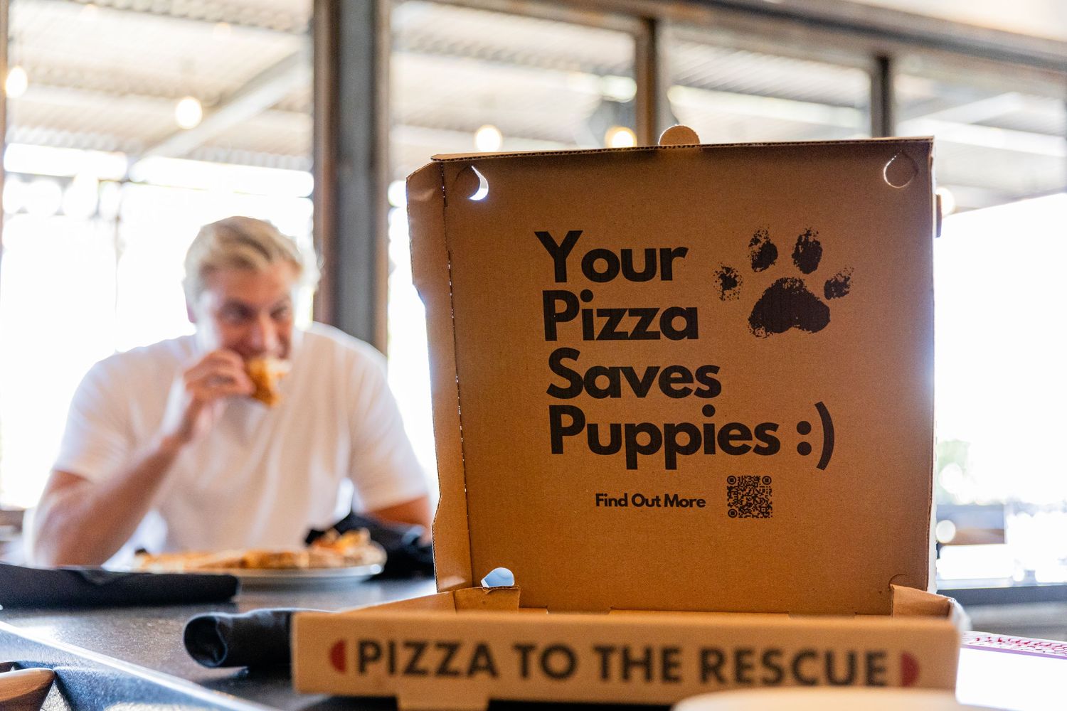 A pizza box reading Your Pizza Saves Puppies with a man eating pizza in the background at a restaurant.