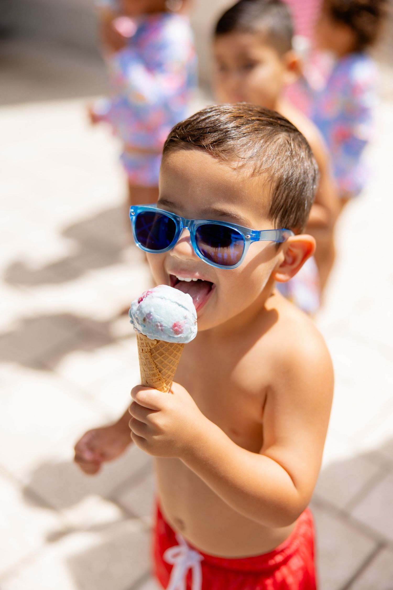 A young boy wearing blue sunglasses and red swim trunks happily licks a large ice cream cone outdoors.