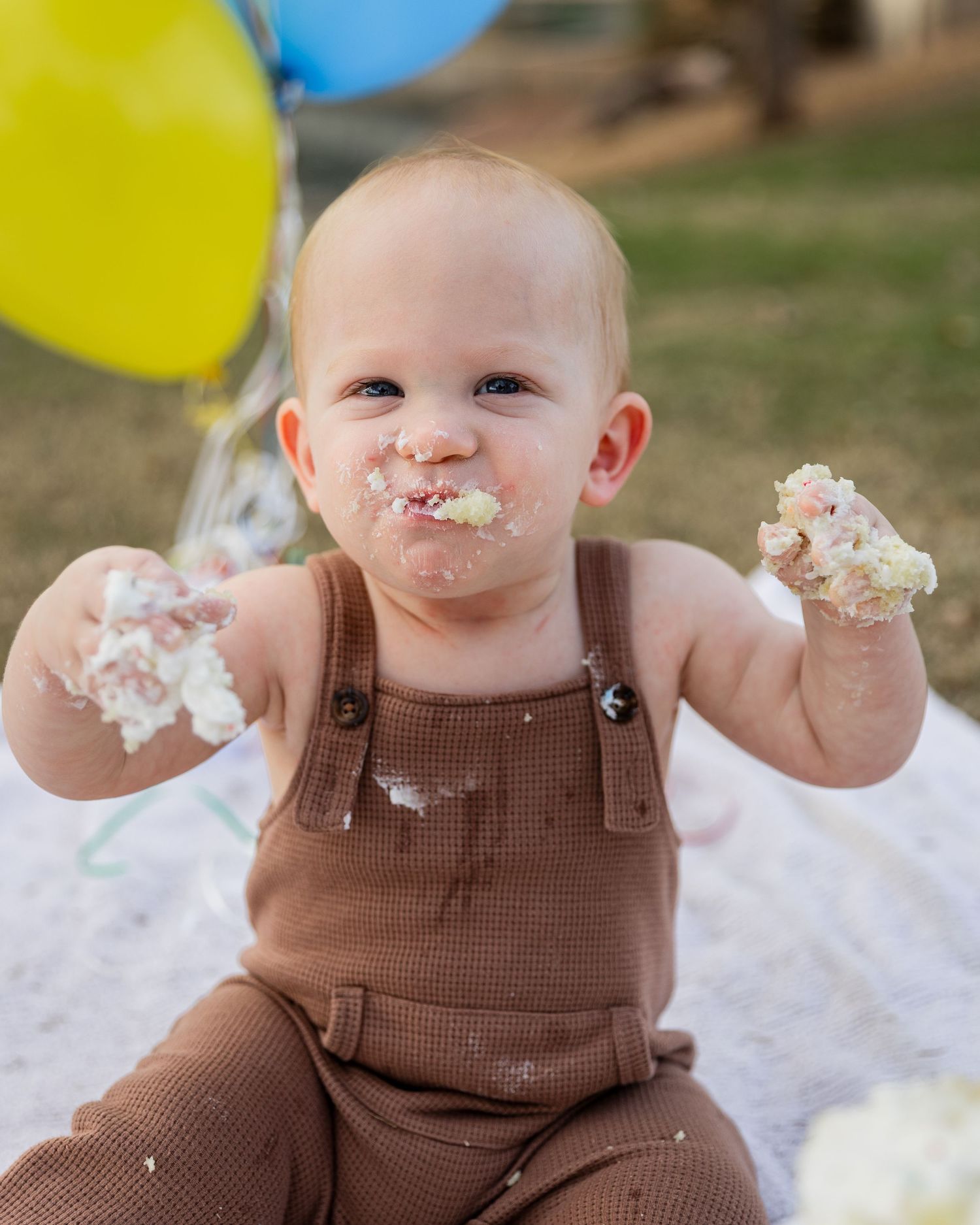 A happy baby with cake-covered face and hands sits outdoors surrounded by colorful balloons at a first birthday.