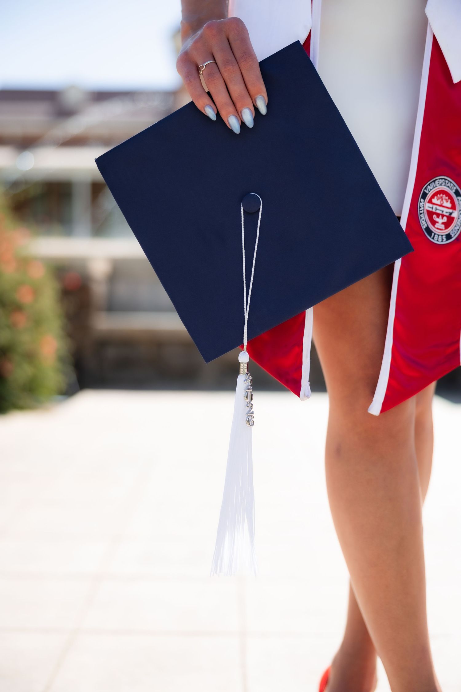 A graduate holds a navy blue mortarboard cap with white tassel, wearing a red academic gown outdoors.