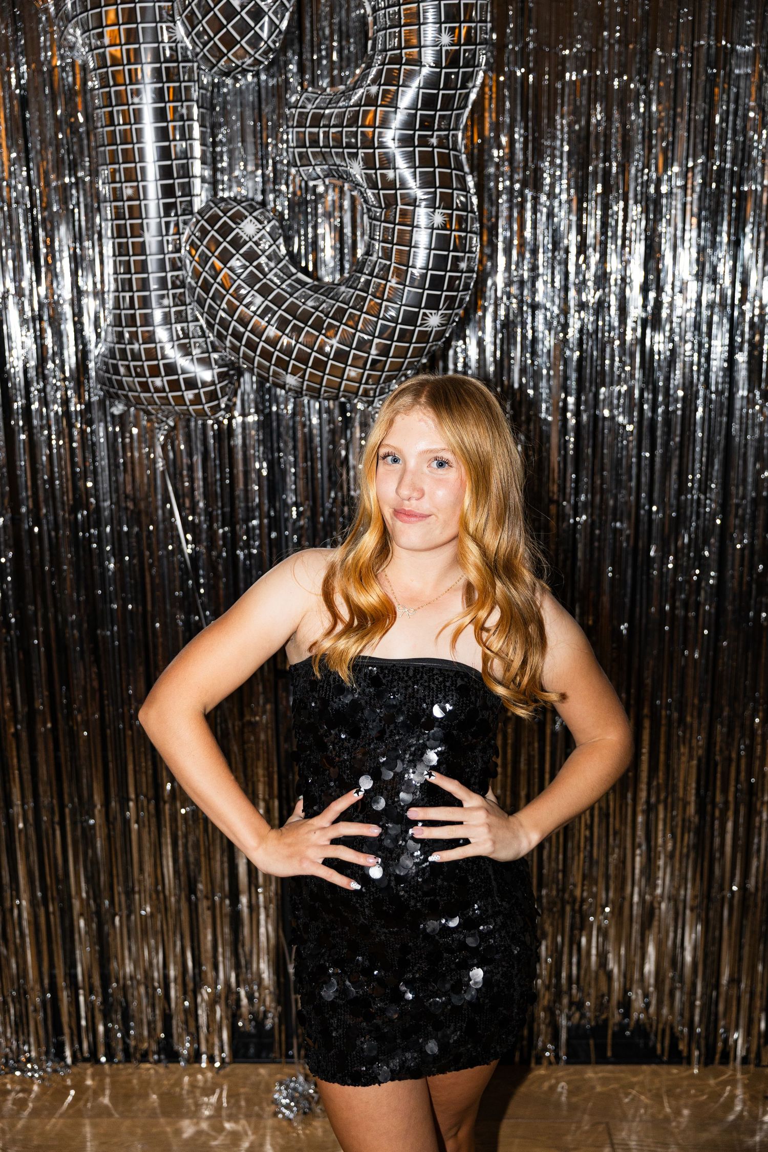 Teen girl in black sequin dress posing in front of silver disco balloon backdrop at a birthday party.