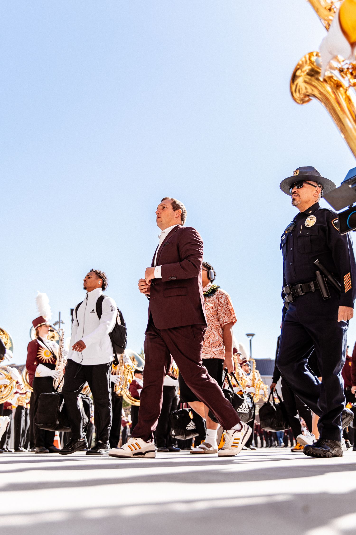 Arizona State University marching band parades outdoors with staff and police escort under bright blue sky.