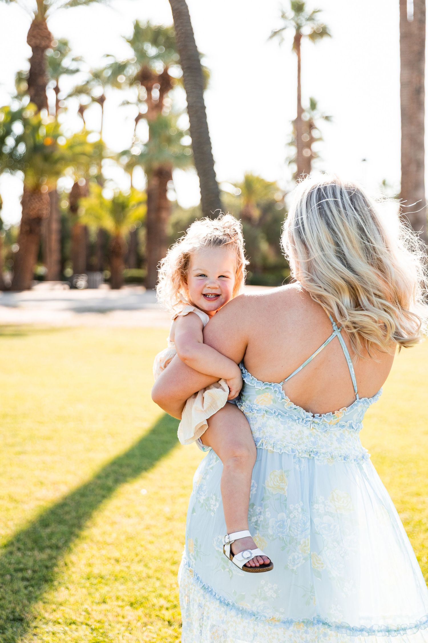 Blonde mother in light blue dress holding smiling toddler in a sunny palm tree park.