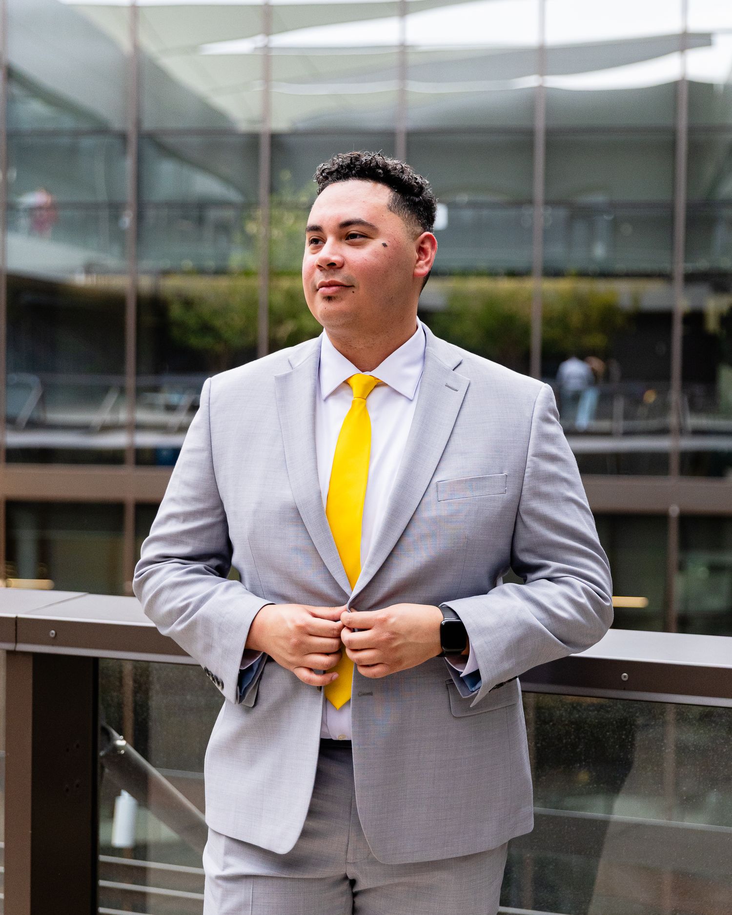 Young man in grey suit with yellow tie adjusting jacket buttons outdoors near glass building.