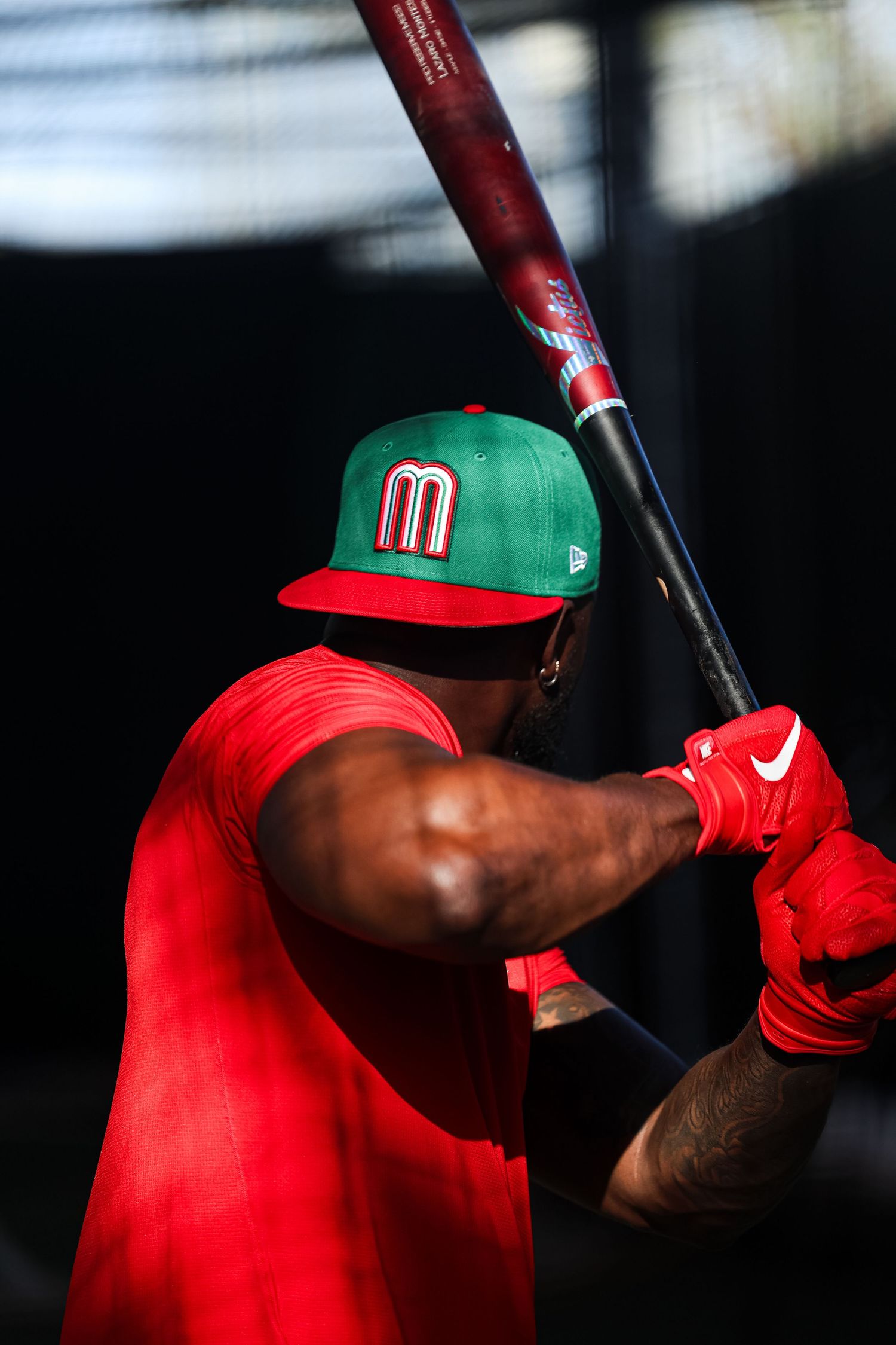 Mexico baseball player in green cap and red jersey takes batting practice in dark stadium setting.