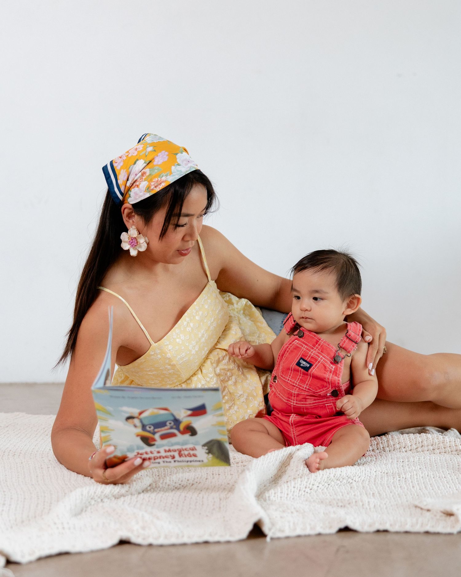 A mother in a yellow dress and floral headband reads a colorful book with her baby in red overalls on a white blanket.