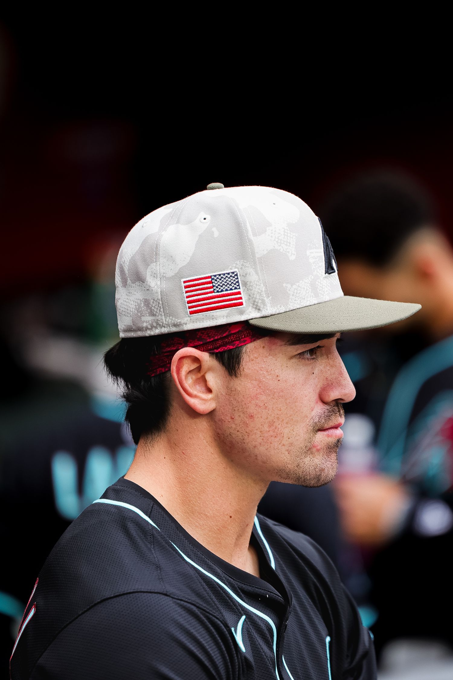 Baseball player wearing American flag cap looks focused in the dugout during a game.