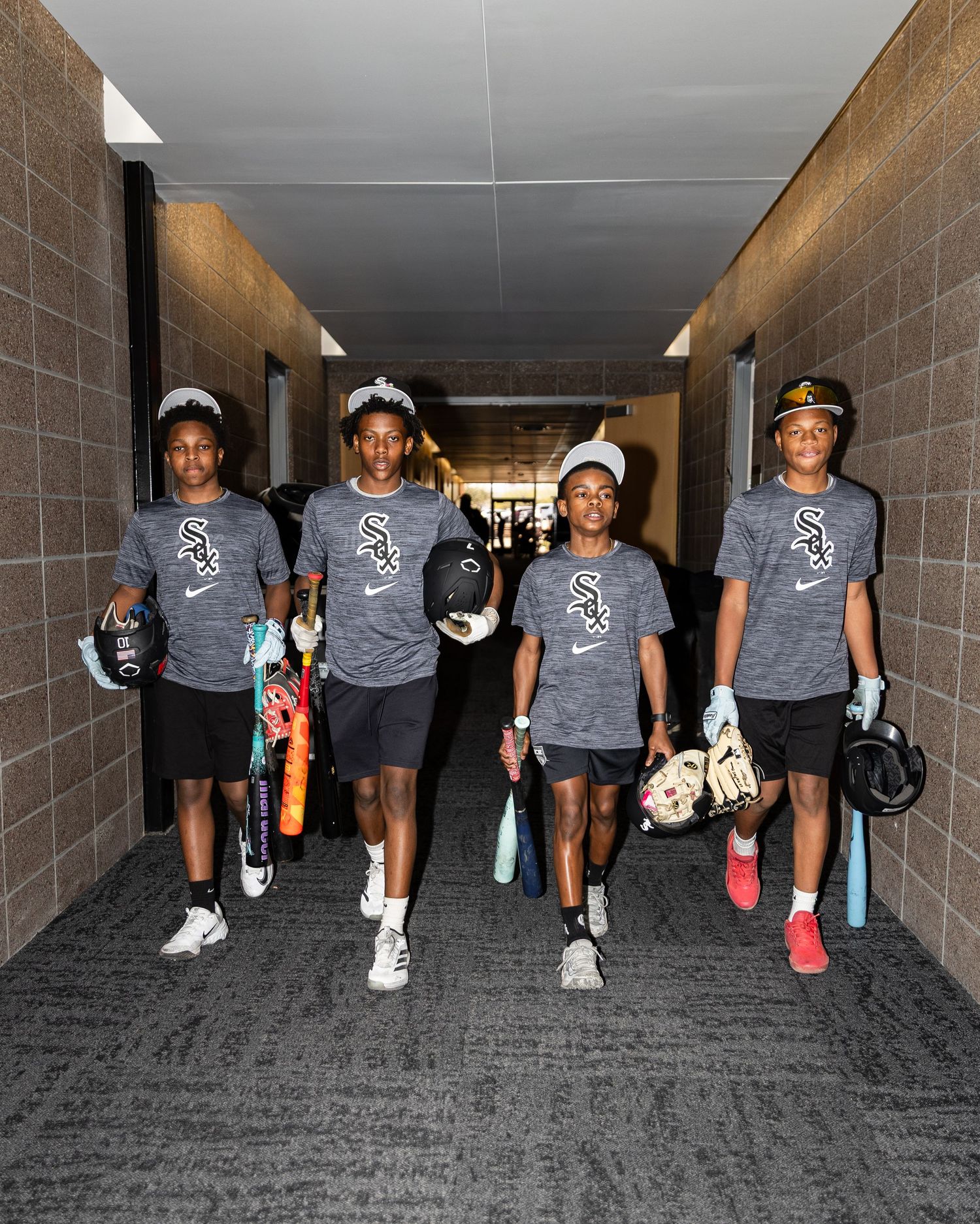 Four young players in Chicago White Sox uniforms walk through a stadium hallway carrying bats and helmets.