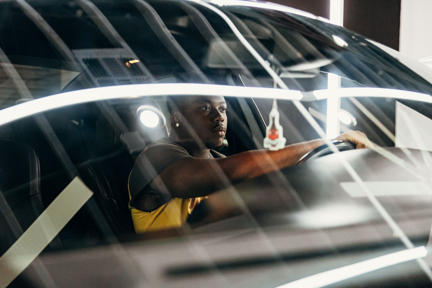 Black man in yellow shirt sitting in a luxury car showroom, viewed through reflective glass windows.