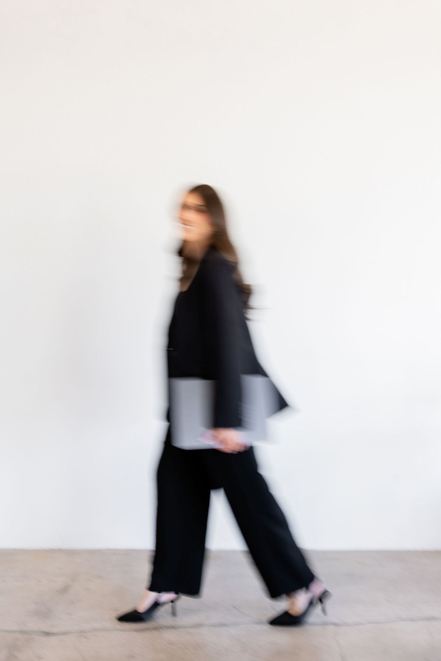 A businesswoman in black suit walks briskly carrying a laptop, captured in motion blur against white wall.