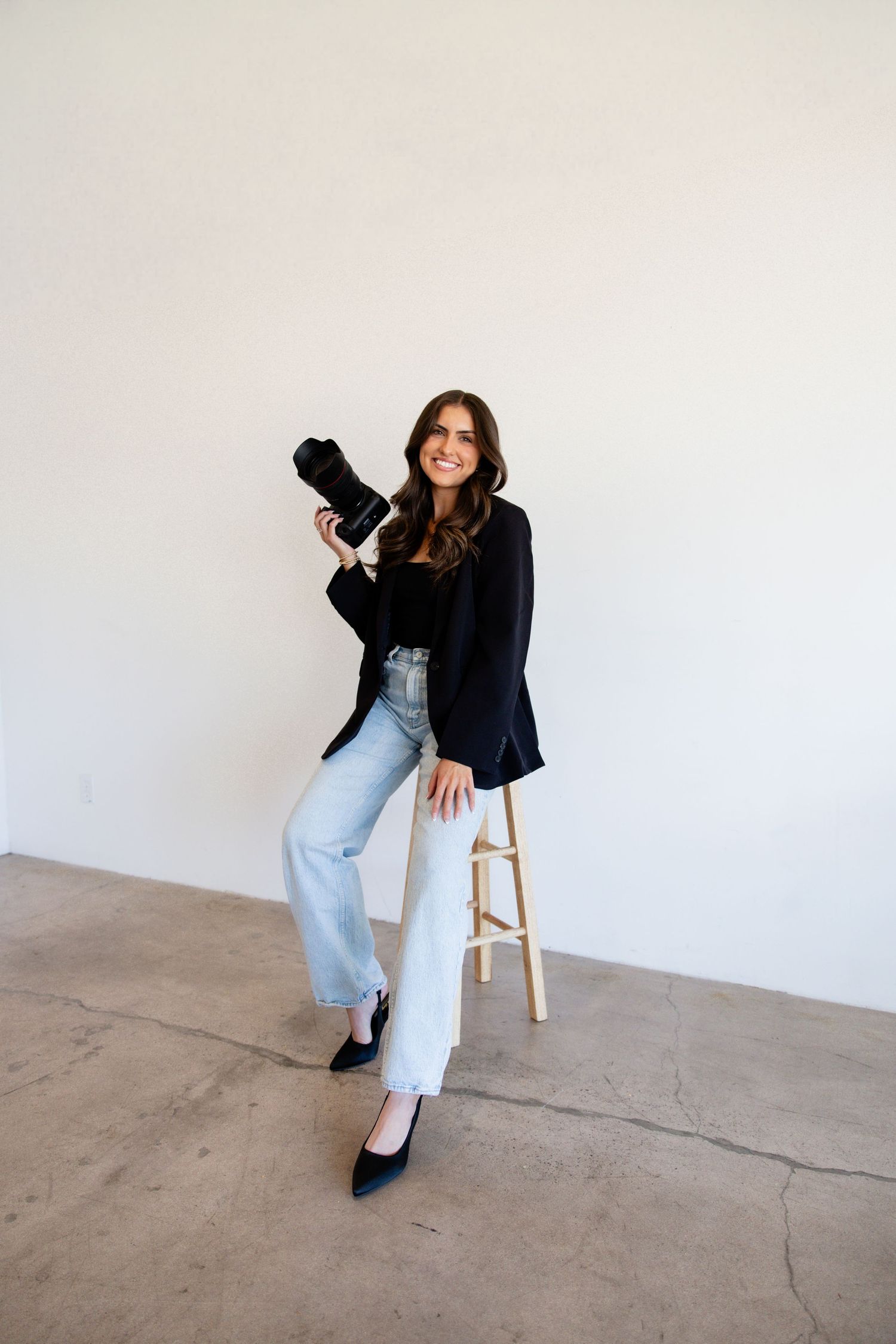 Smiling female photographer in black blazer and jeans posing with DSLR camera on wooden stool.