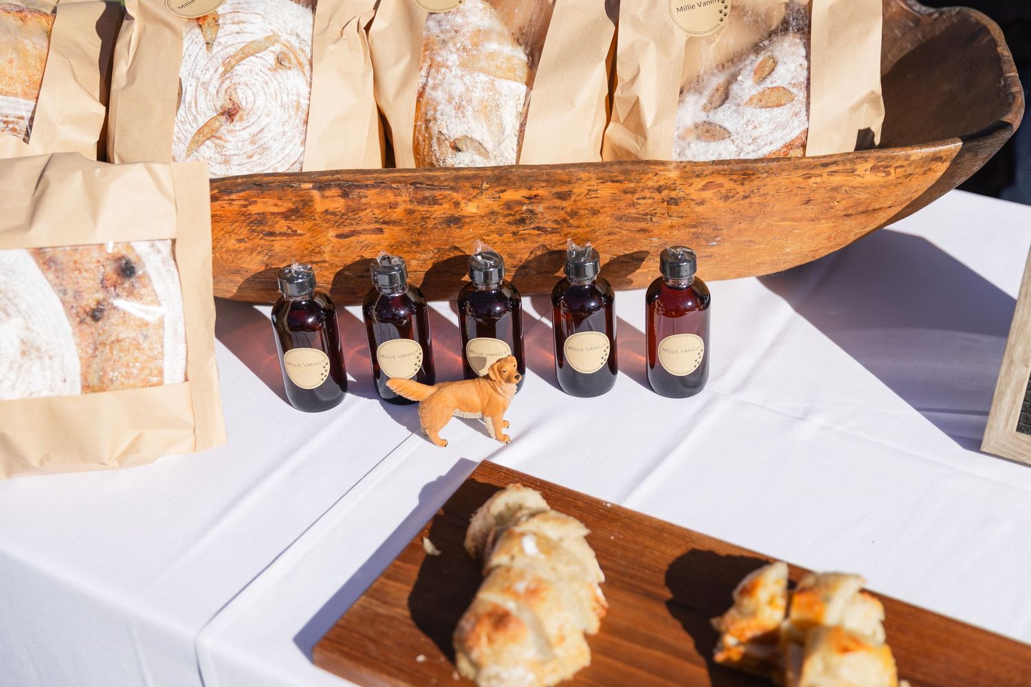 Row of dark glass bottles with bread loaves on wooden board at outdoor artisan food vendor table.