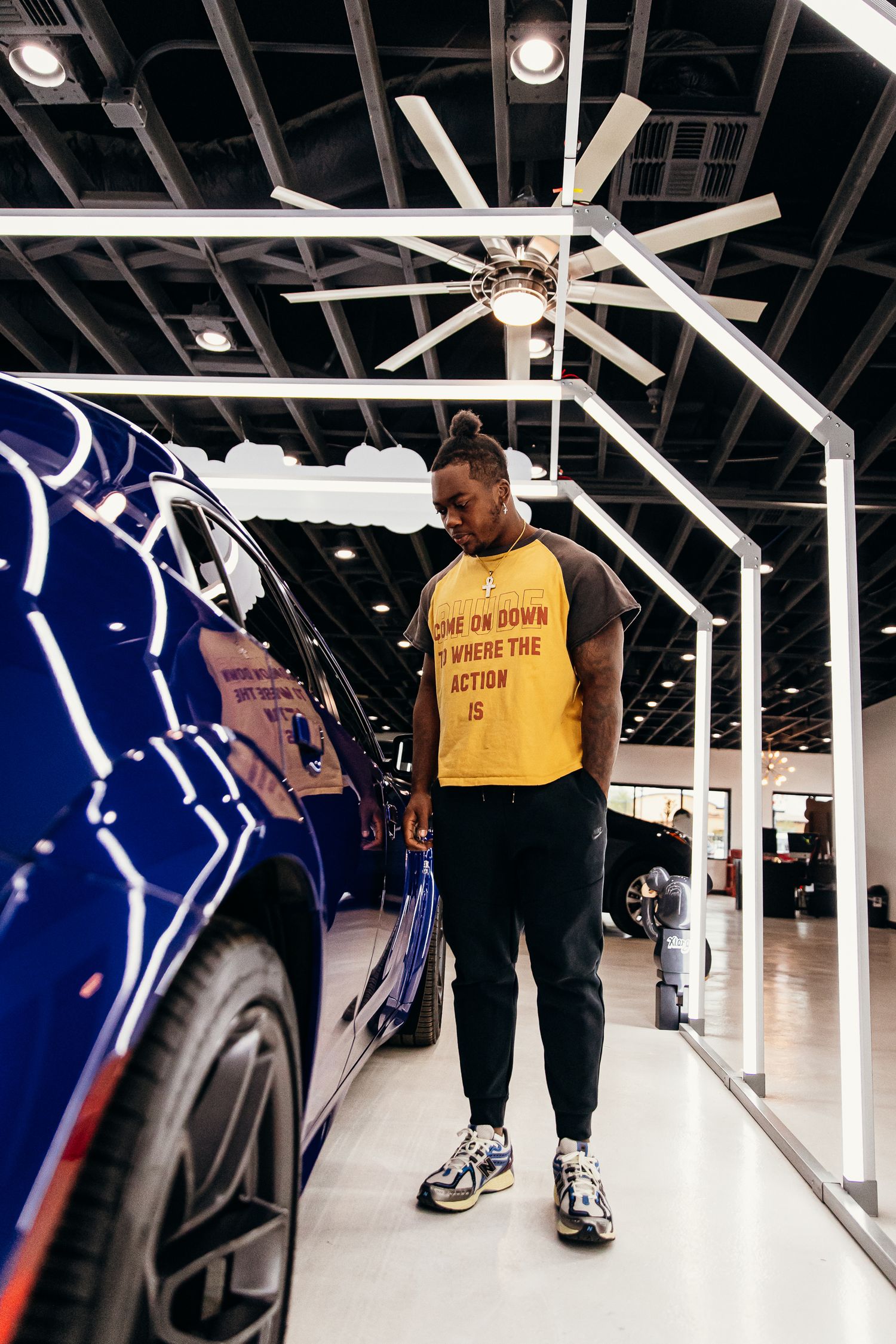 Man in yellow graphic tee and black joggers standing in a car showroom beside a blue vehicle.