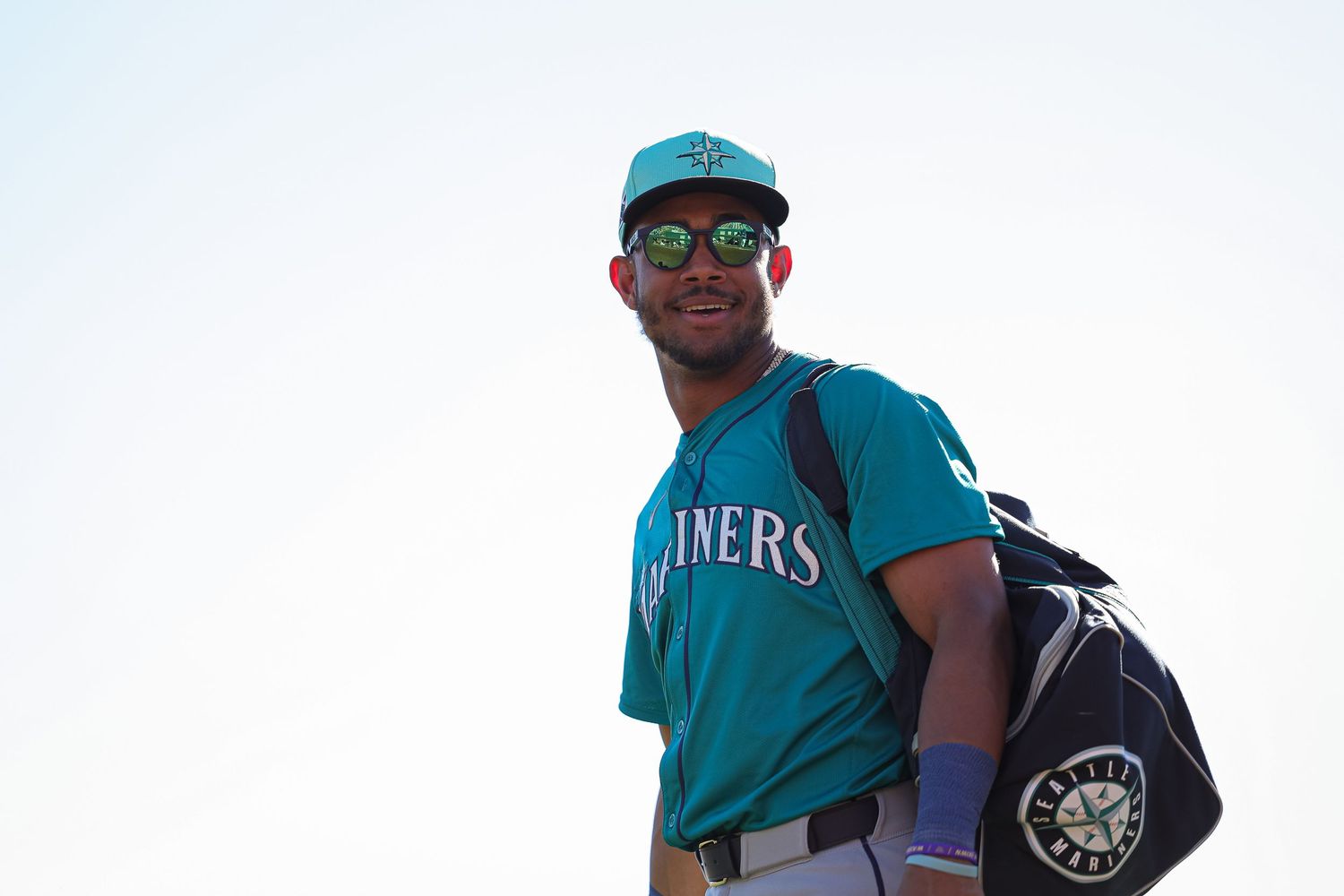 Seattle Mariners player smiling with green sunglasses and team duffel bag during spring training.