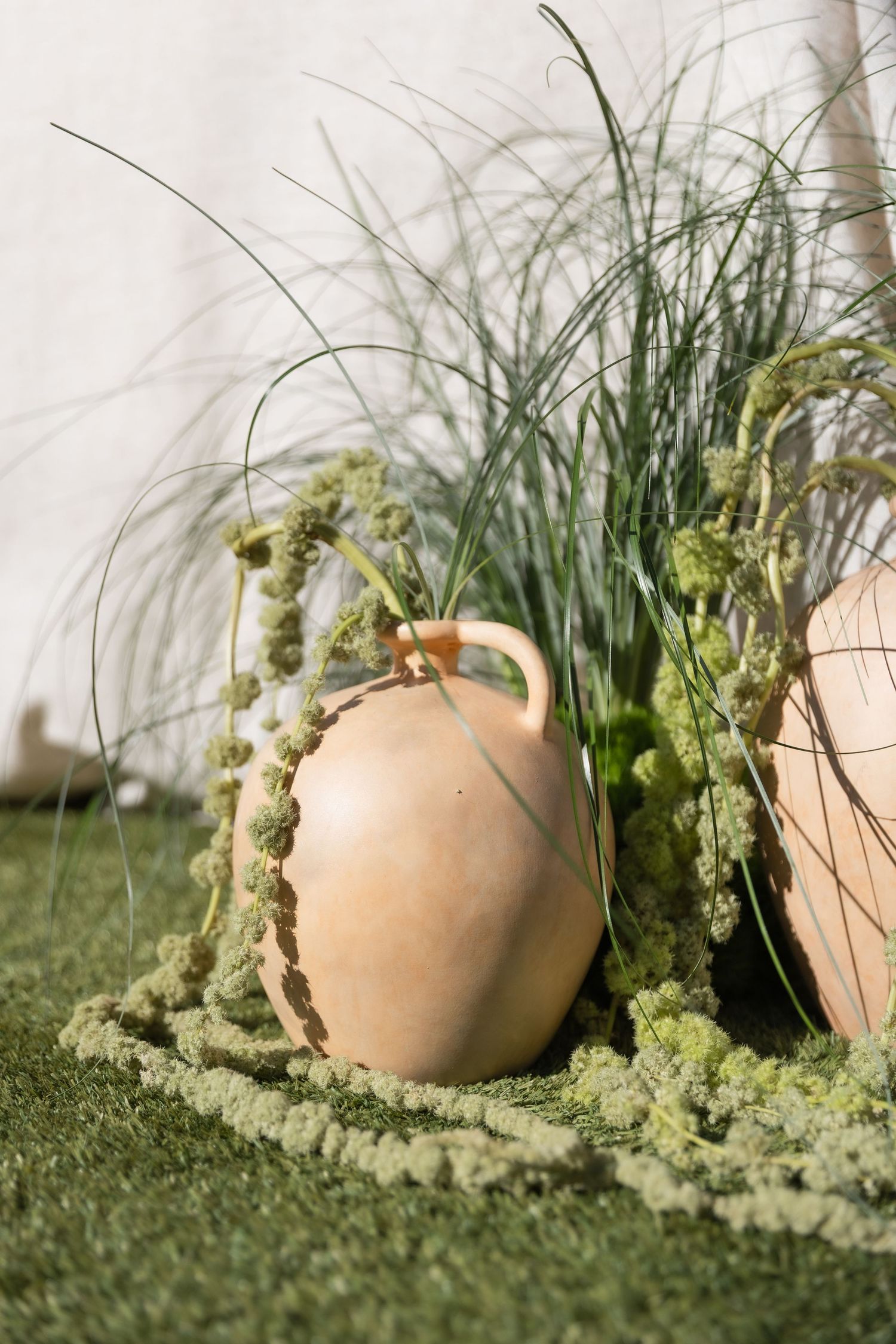 Rustic terracotta jugs surrounded by green moss and ornamental grass on artificial turf.