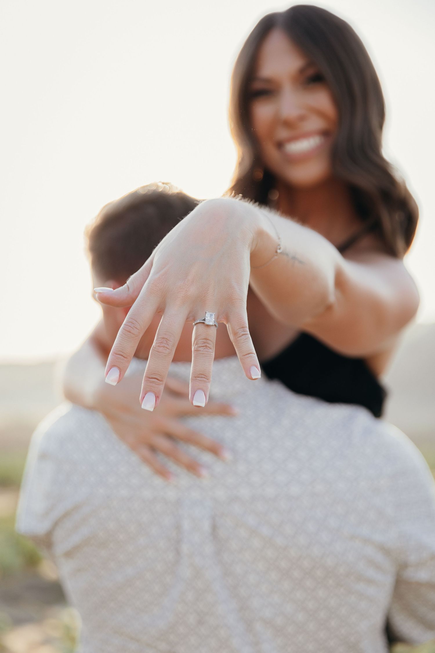 Smiling woman hugging man while showing off her diamond engagement ring during an outdoor proposal photo session.