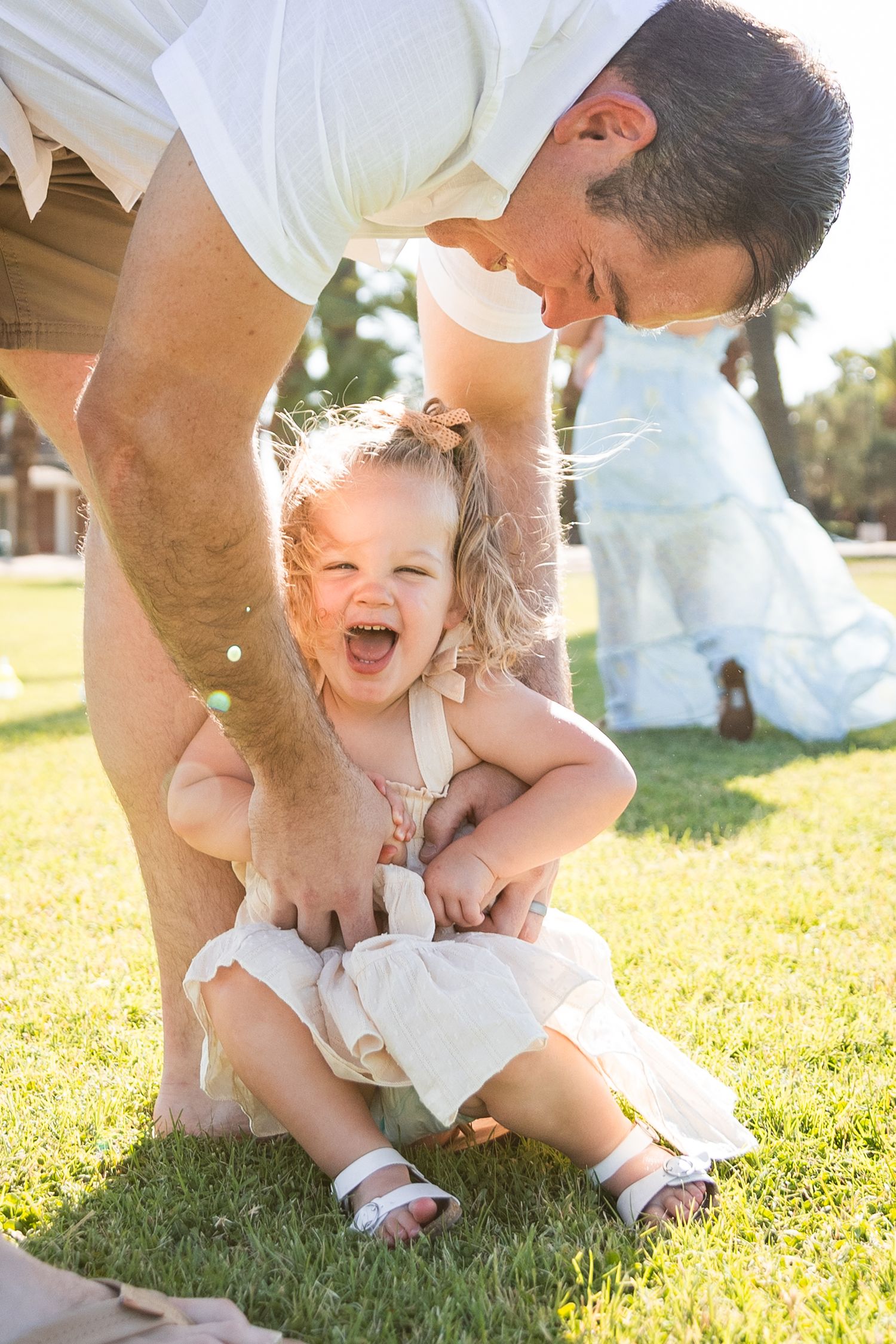 Laughing toddler girl in white dress being held by father on sunny green grass park lawn.