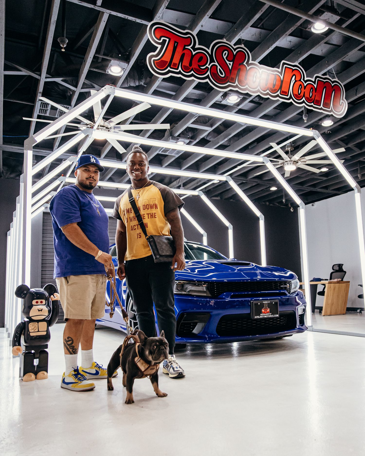 Two men and a dog pose with a blue Dodge Charger inside The Showroom car dealership with neon lights.
