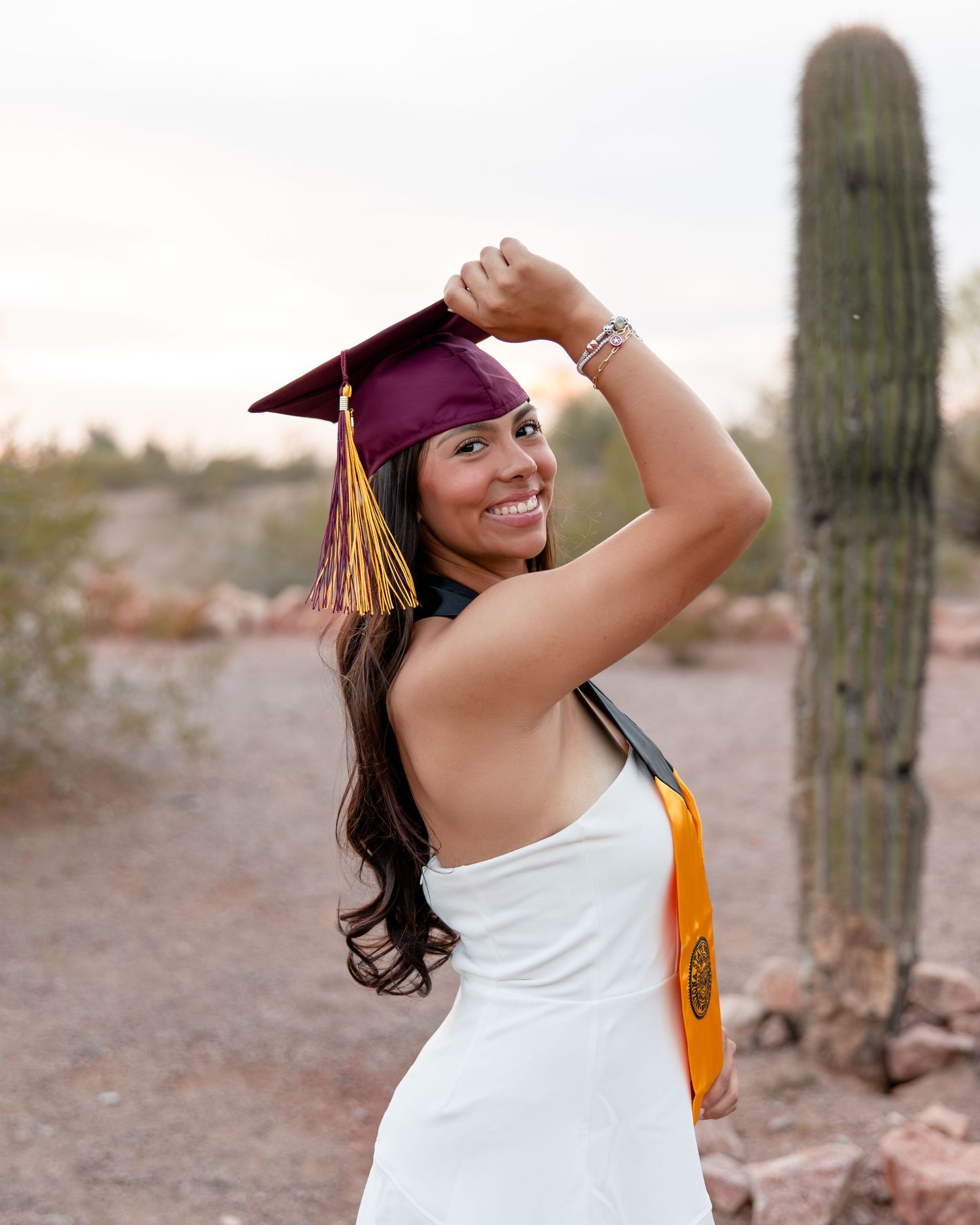 A smiling graduate in a white dress adjusts her maroon ASU cap beside a tall saguaro cactus at sunset.