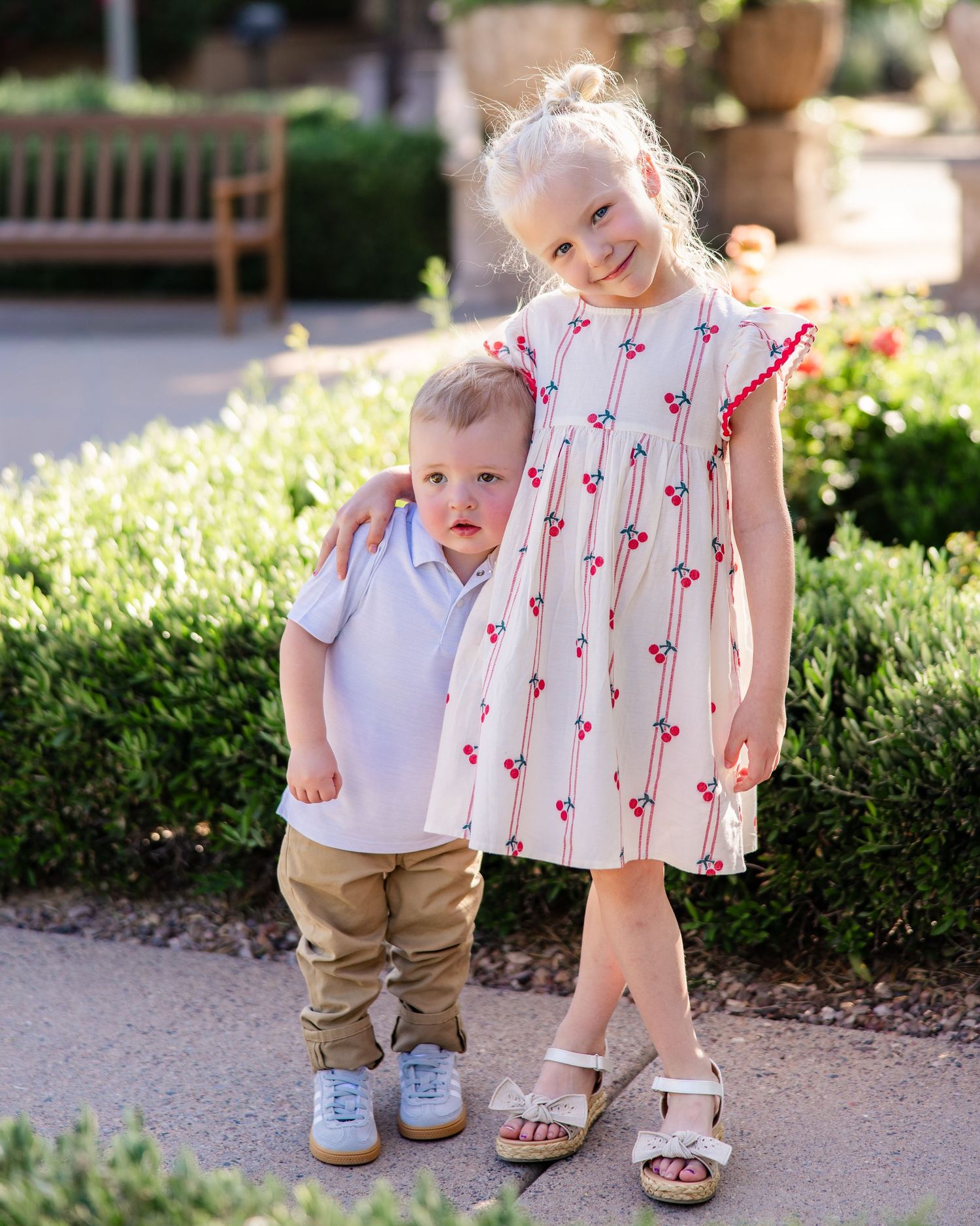 Young girl in cherry print dress with arm around toddler boy in white polo outdoors in garden setting.