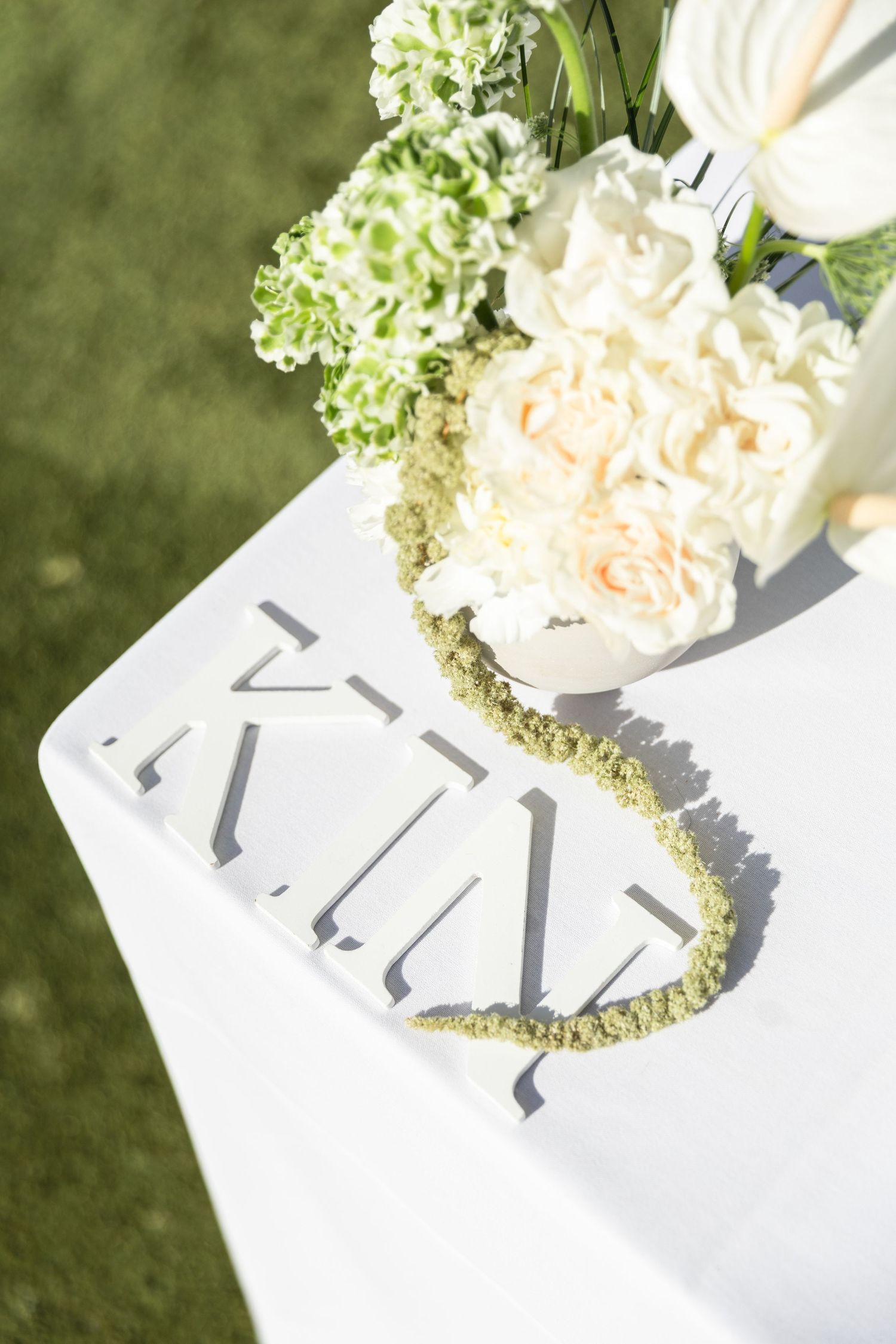 Elegant white floral centerpiece with green wreath on a white tablecloth at an outdoor event.