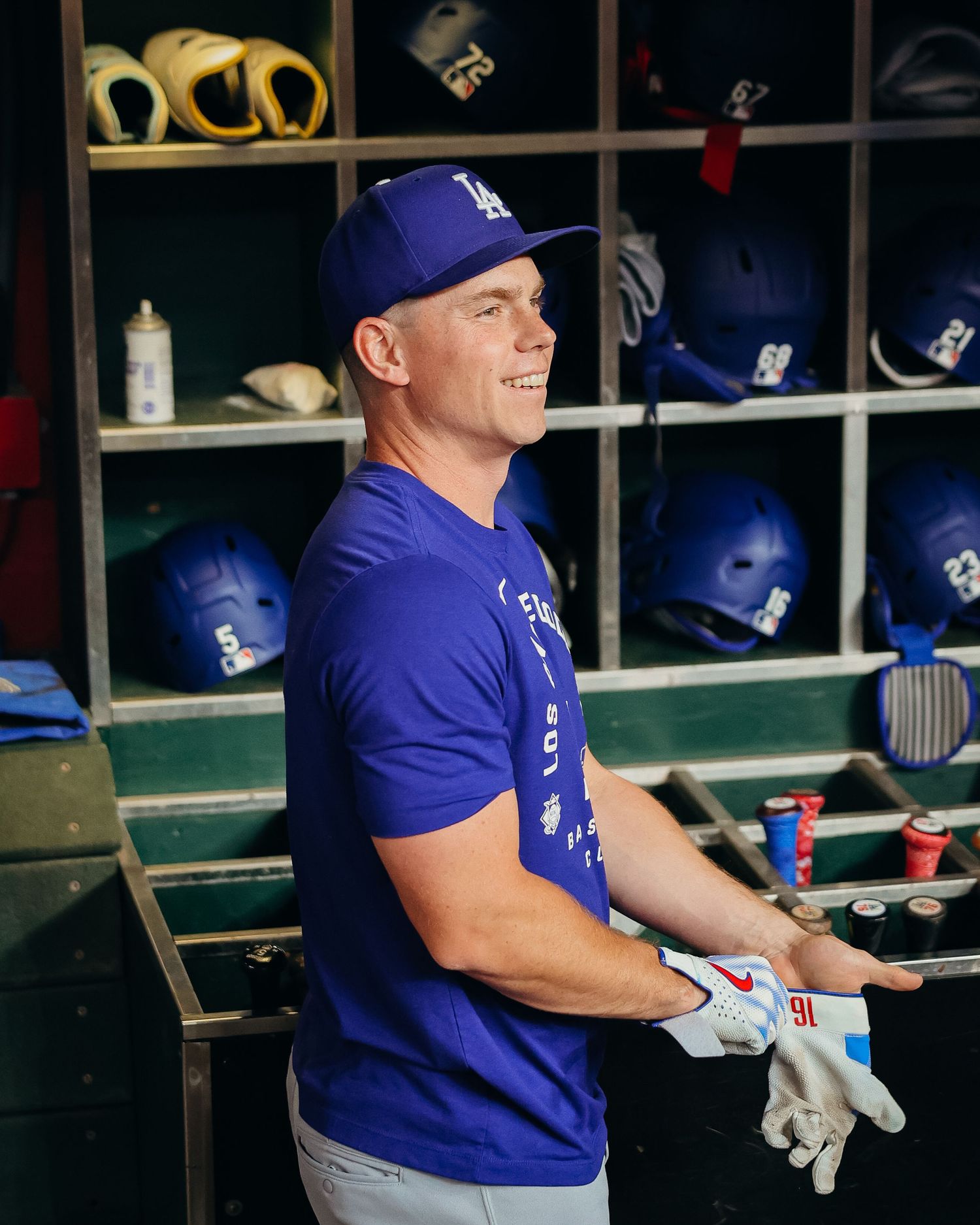 Los Angeles Dodgers player smiles in the dugout wearing a blue team t-shirt and white batting gloves.