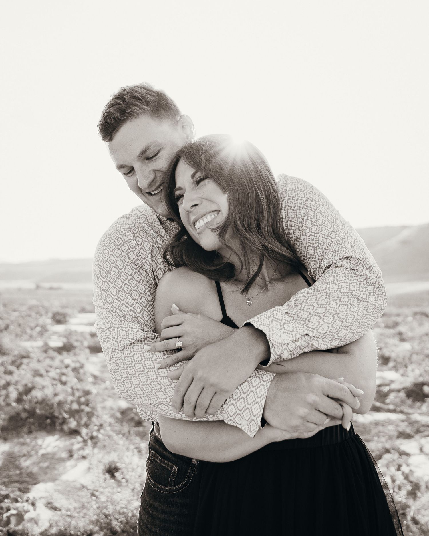 Black and white photo of a couple embracing outdoors, woman laughing joyfully as man hugs her from behind in sunny landscape.