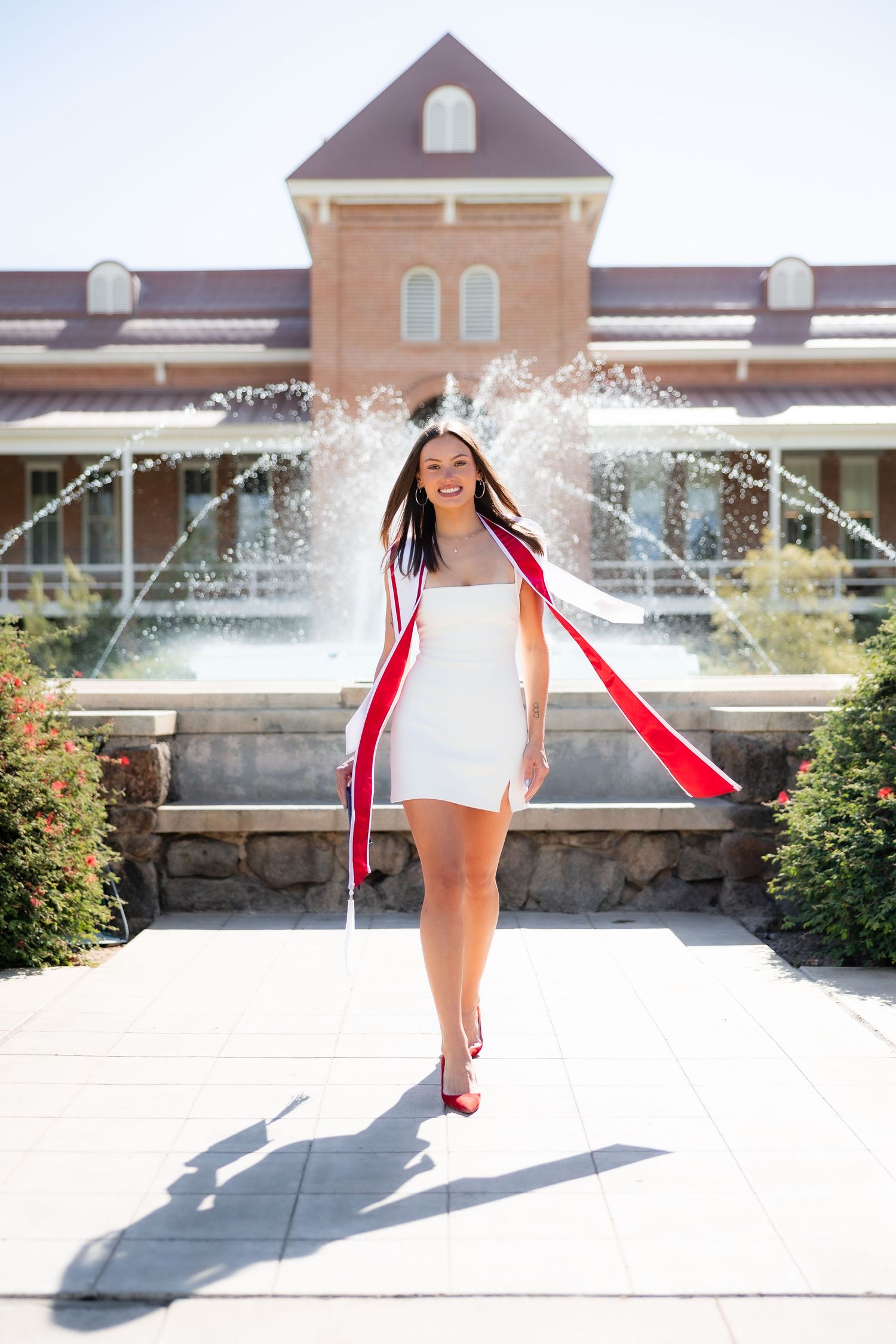 Smiling graduate in white dress and red academic regalia poses before a fountain outside a brick university building.