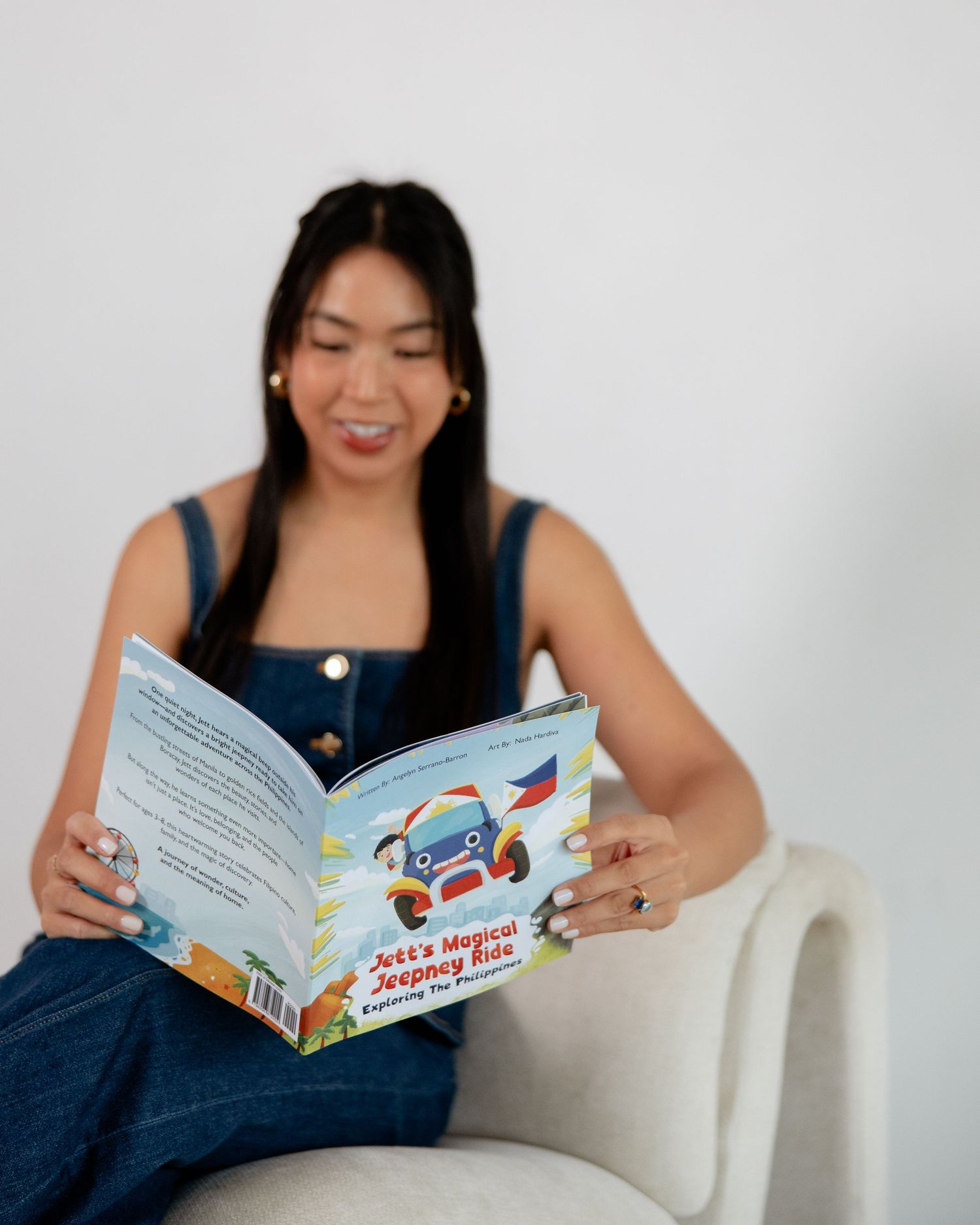 Woman smiling while reading Jets Magical Jeepney Ride childrens book seated in white chair wearing denim overalls.