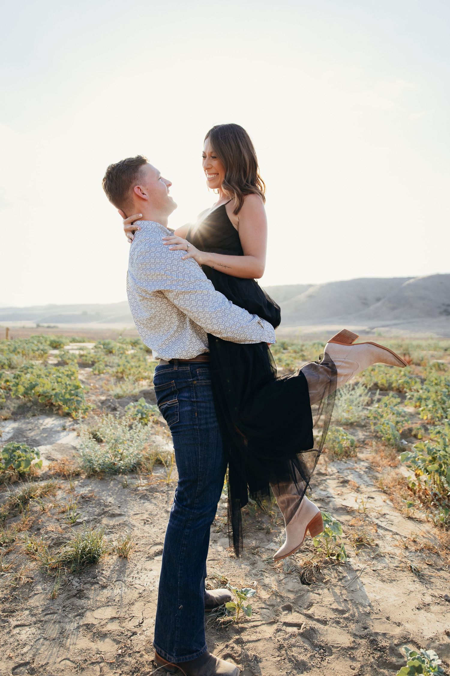 Man lifting woman in his arms in a romantic desert landscape, both smiling and gazing at each other under soft sunlight.