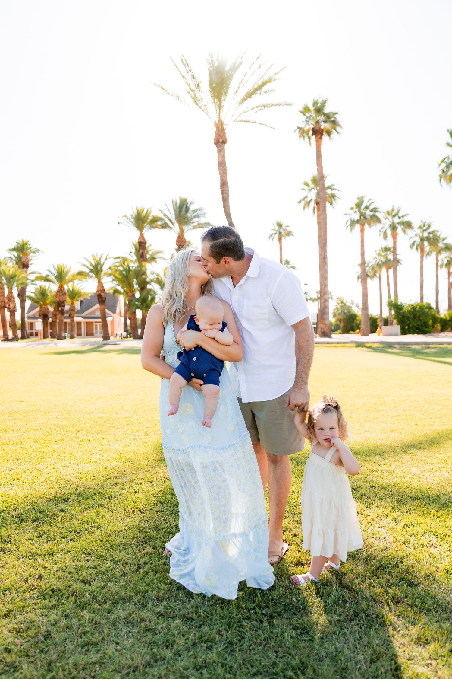 Family of four kissing and smiling on green lawn surrounded by tall palm trees at sunset.