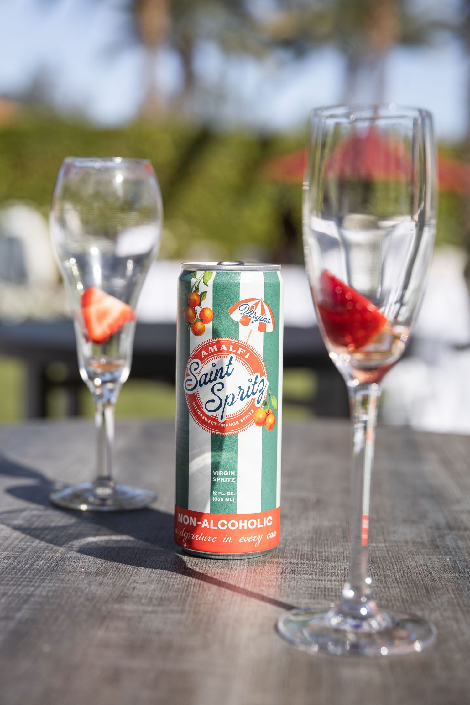 A can of Amalfi Saint Spritz non-alcoholic spritz beside two champagne glasses with strawberries outdoors.