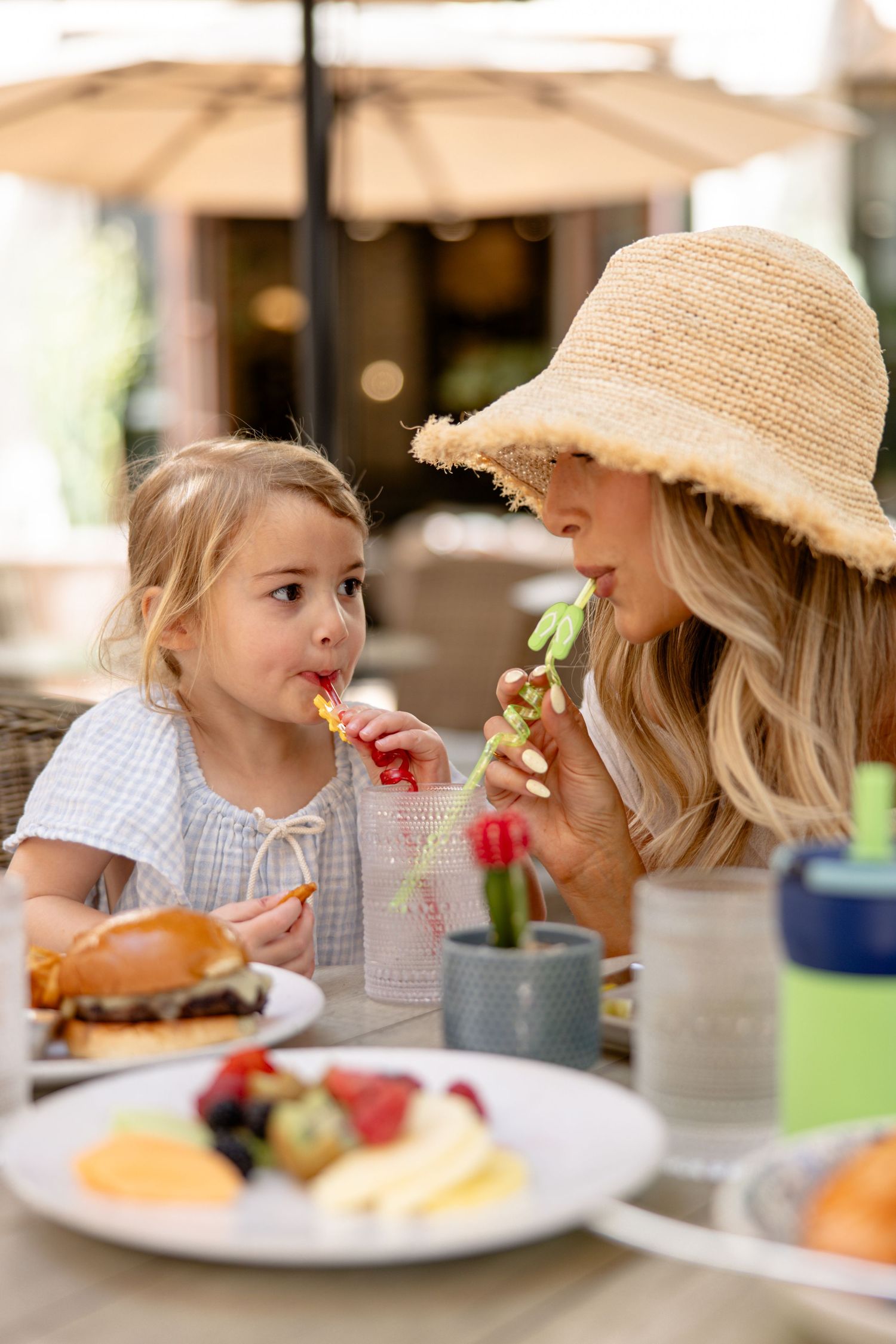 A mother and young daughter share drinks through colorful straws at an outdoor restaurant table filled with food.