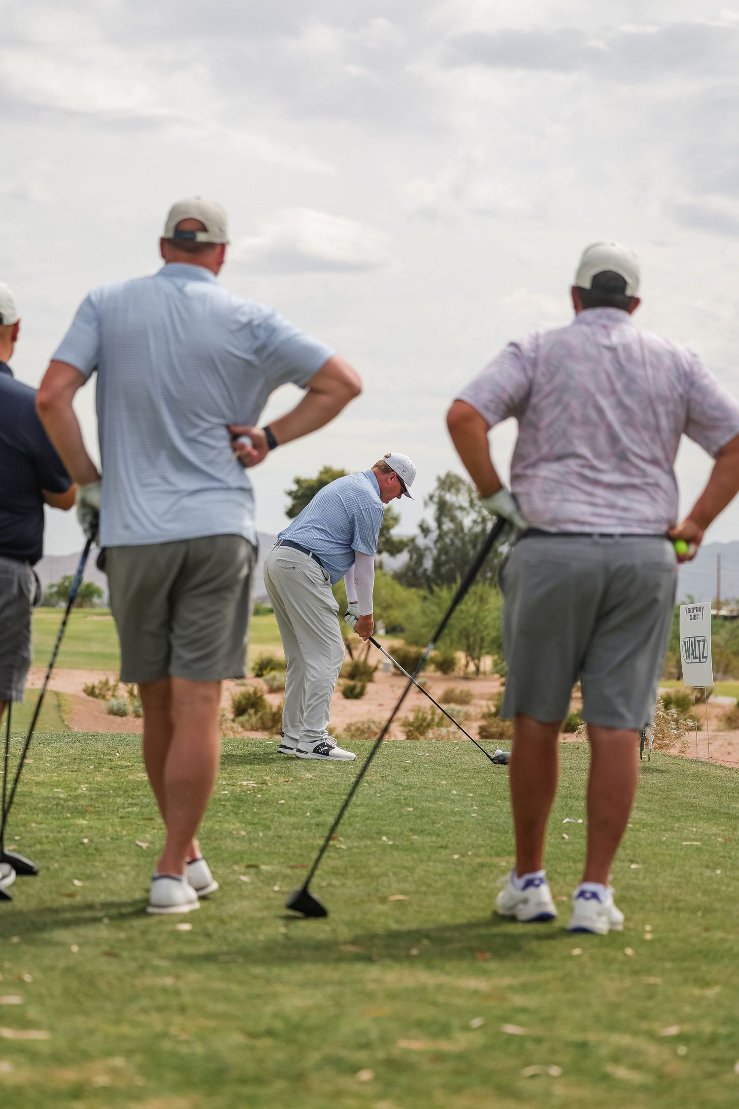 Four golfers on a golf course tee box, one player preparing to swing while others watch.