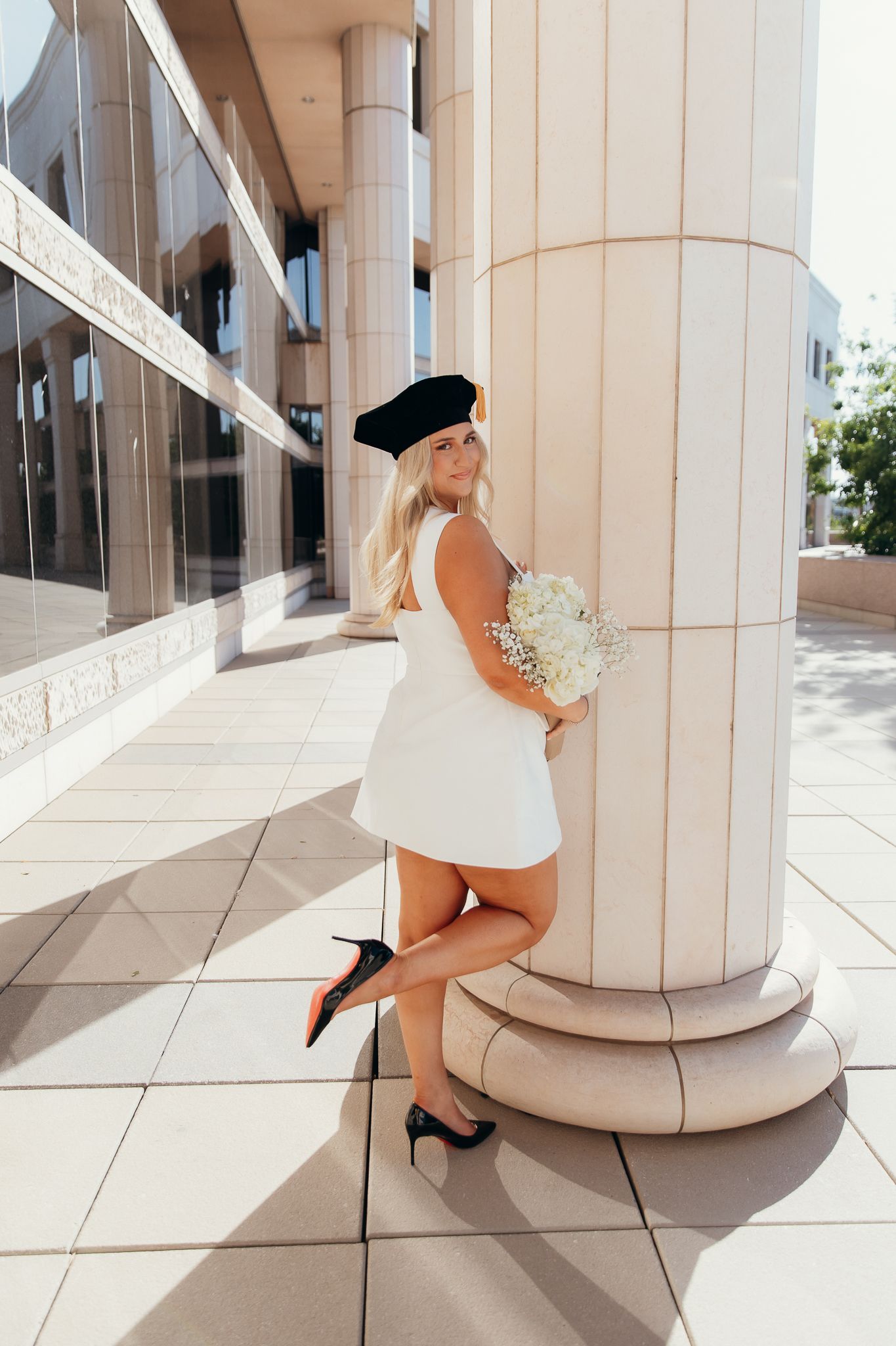 Elegant blonde woman in white mini dress and black beret leans against column holding flowers.