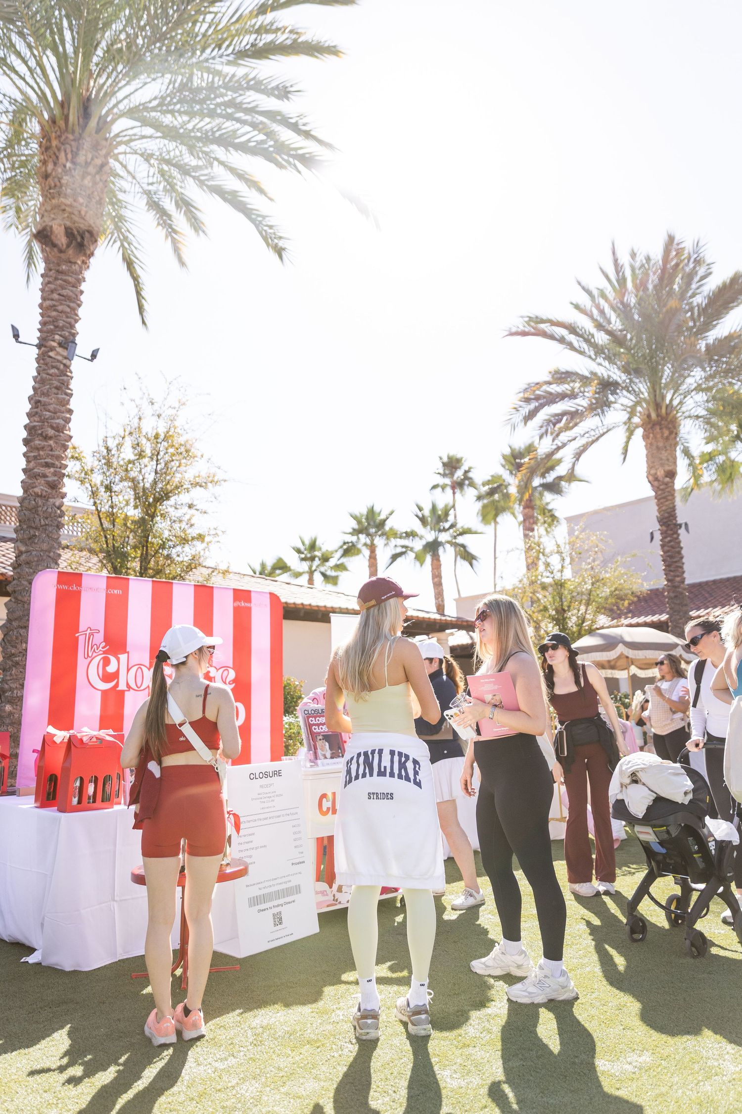 Women gathered at a red and white striped booth under tall palm trees outdoors.