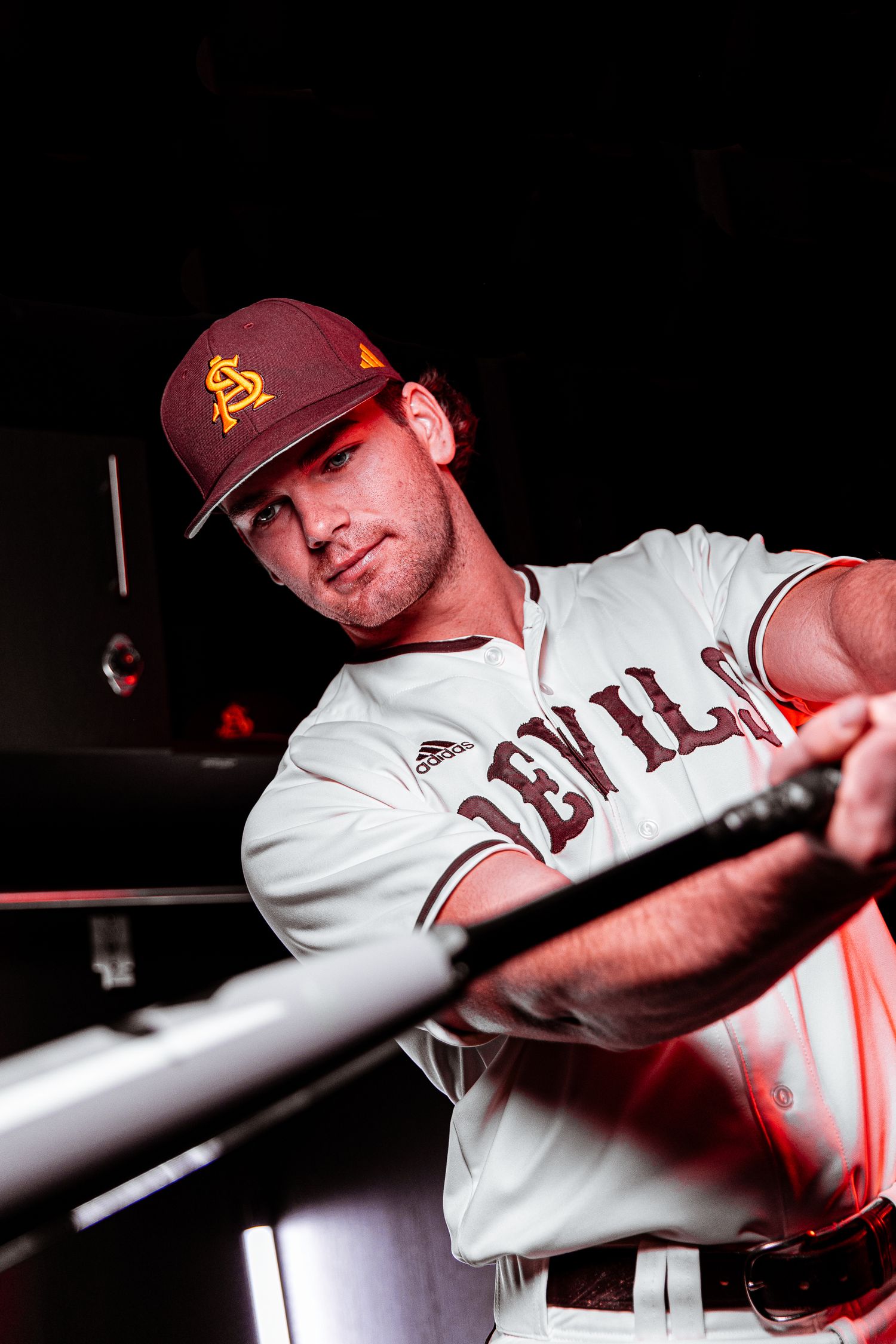 Arizona State Sun Devils baseball player leans on dugout rail holding bat in white Devils uniform.