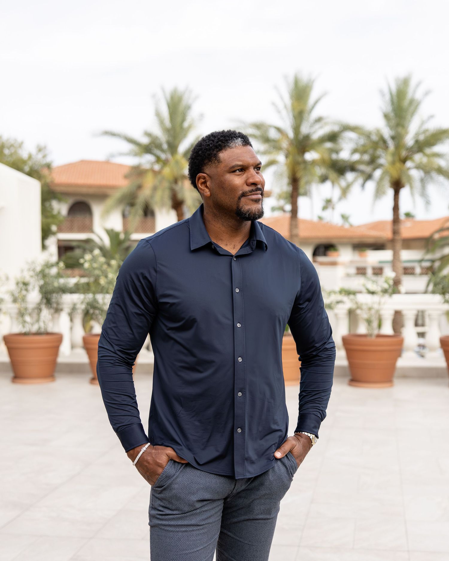 Confident Black man in navy shirt with hands in pockets posing outdoors near palm trees and resort building.
