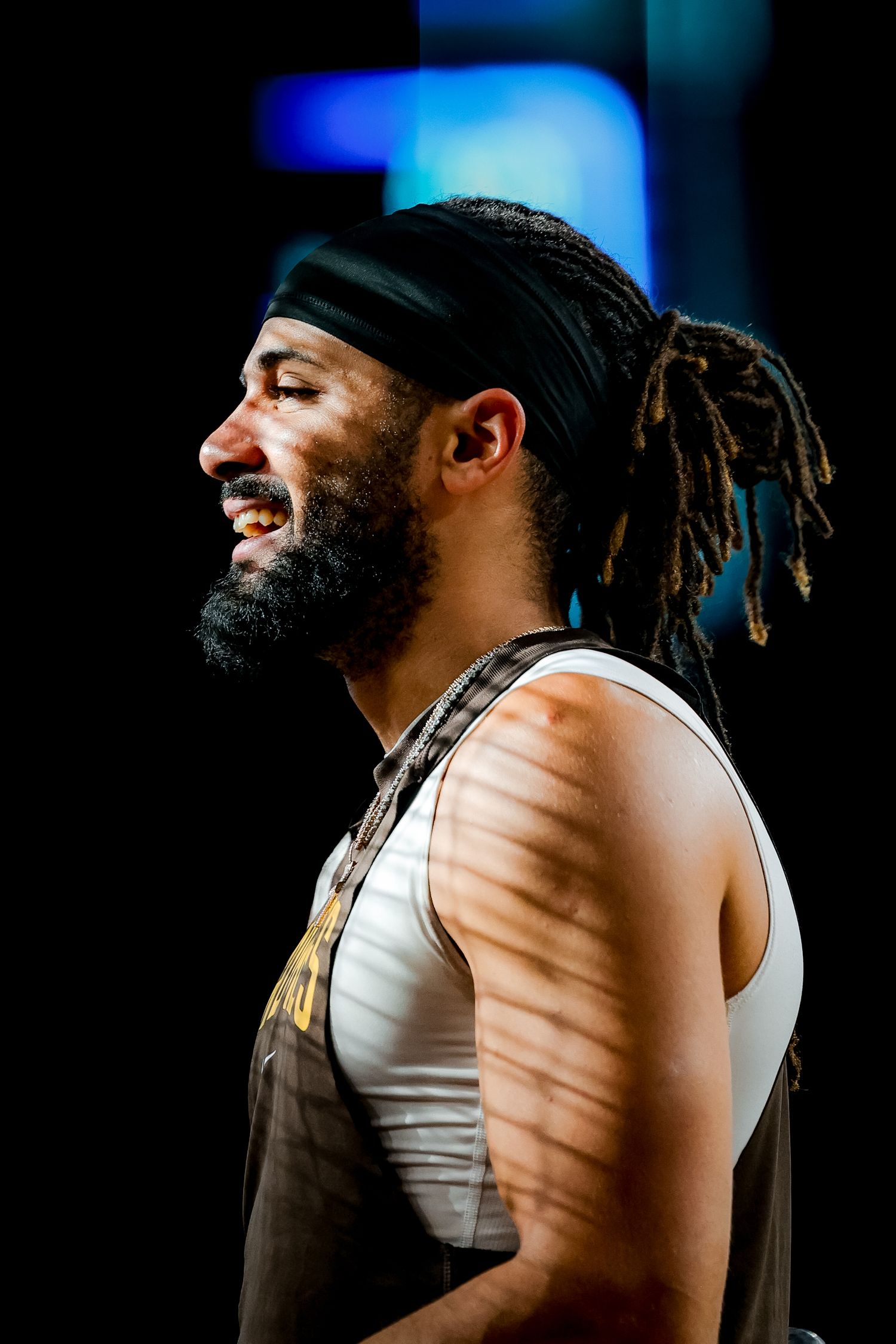 Smiling bearded athlete with dreadlocks and headband poses in dramatic side lighting.