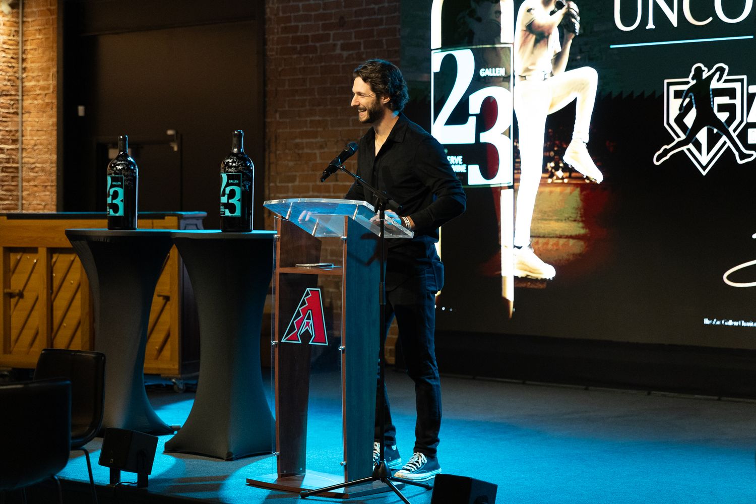 Man speaking at podium during Uncork for a Cause charity event with Arizona Diamondbacks logo displayed.