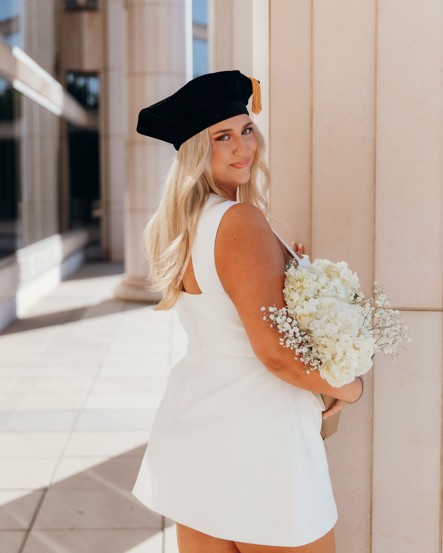 Blonde woman in white dress and black graduation cap holding white flowers.