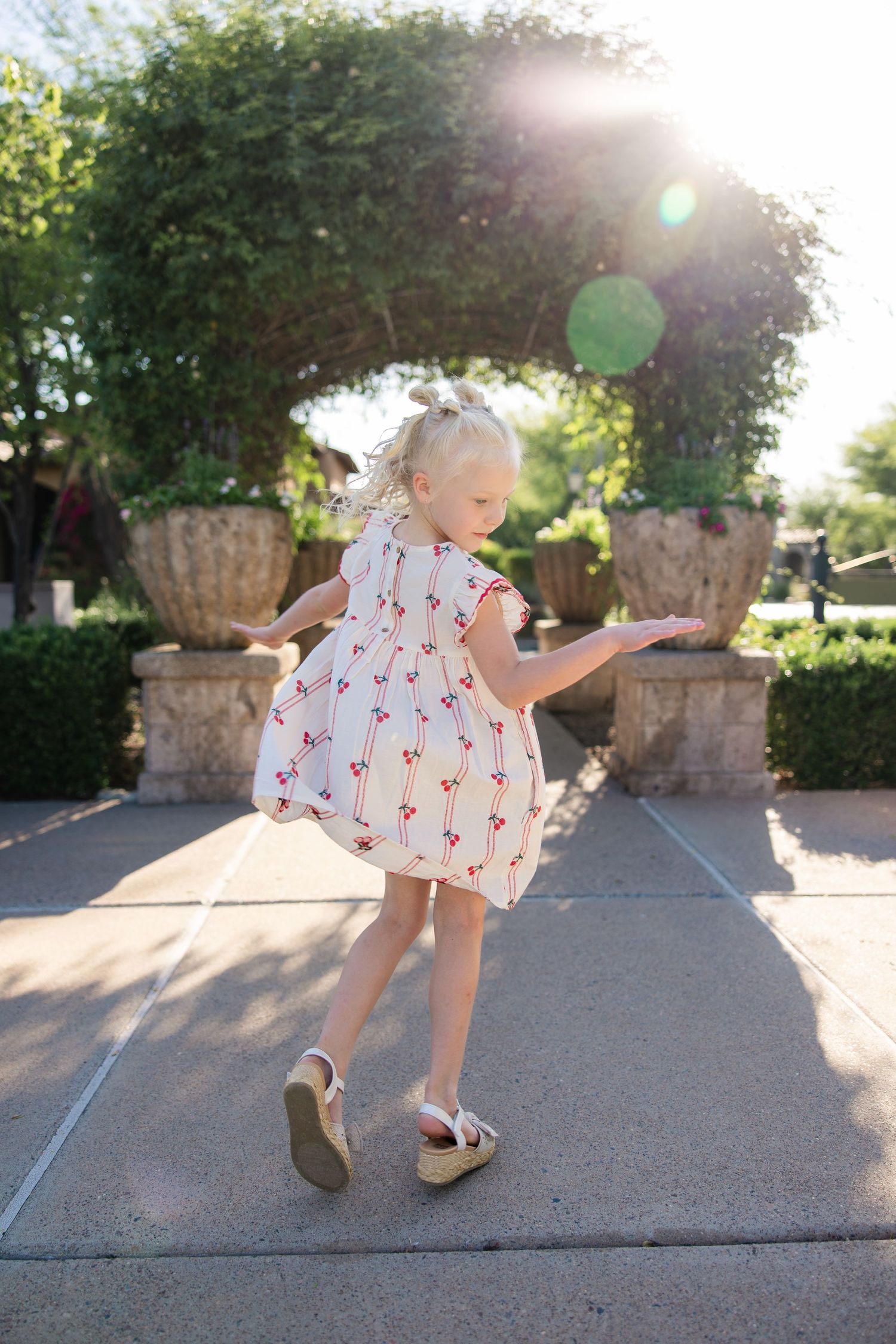 Blonde toddler girl in cherry print dress twirling outdoors in sunny garden setting.