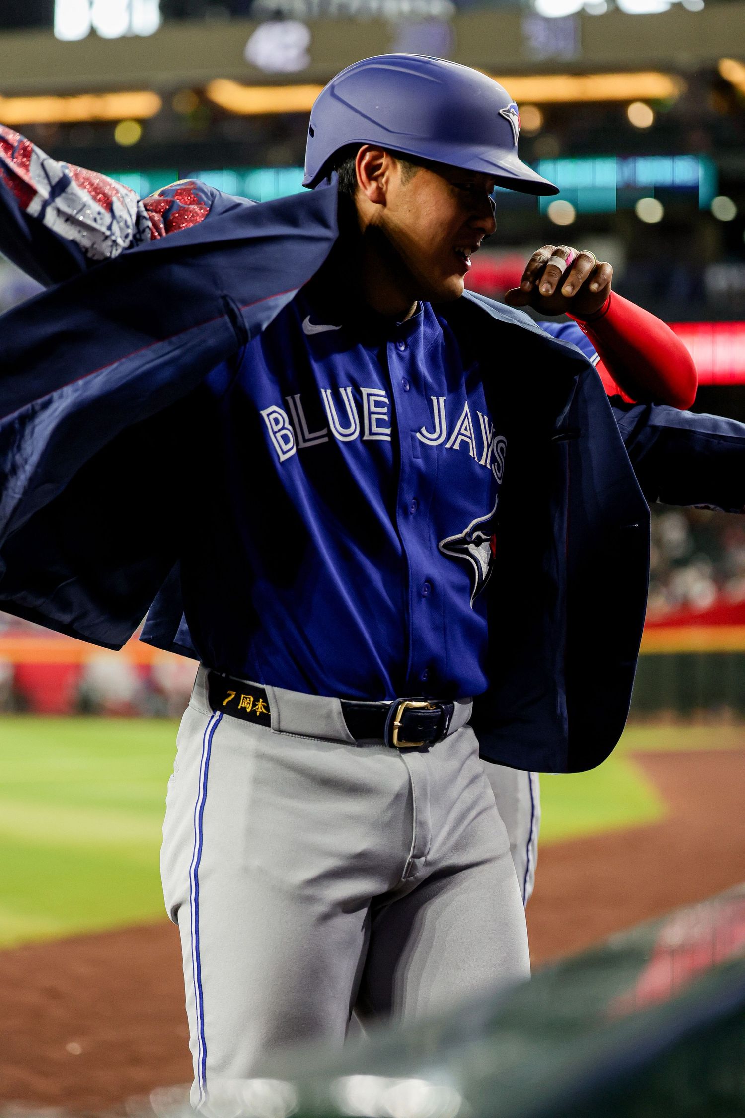 Toronto Blue Jays player putting on a navy jacket over his uniform while wearing a batting helmet in the dugout.