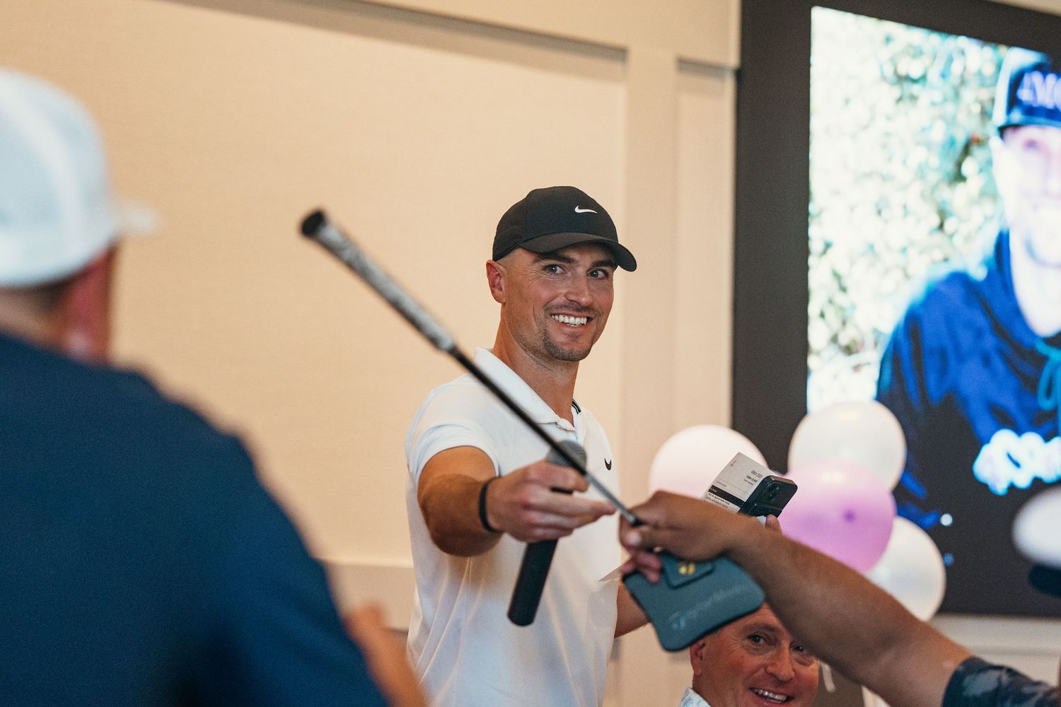 Smiling golfer handing a signed club to a fan at an indoor charity golf event signing.