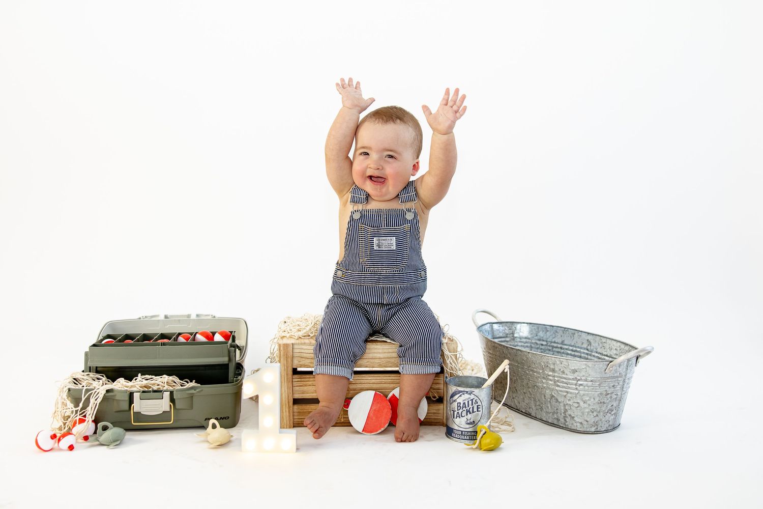 Happy baby in denim overalls sitting on a wooden crate with arms raised, surrounded by vintage toys.