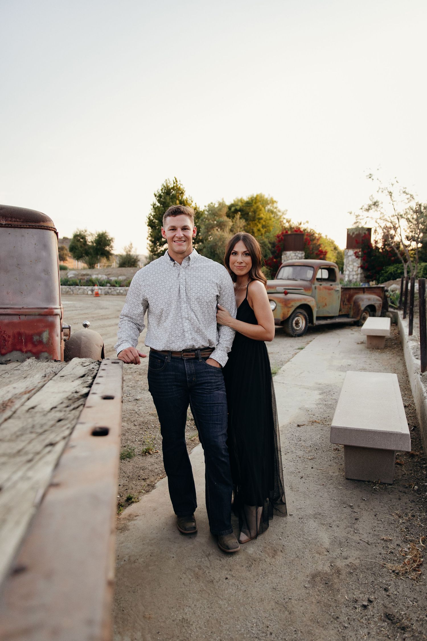 Couple posing together outdoors near rustic vintage trucks at golden hour on a farm setting.